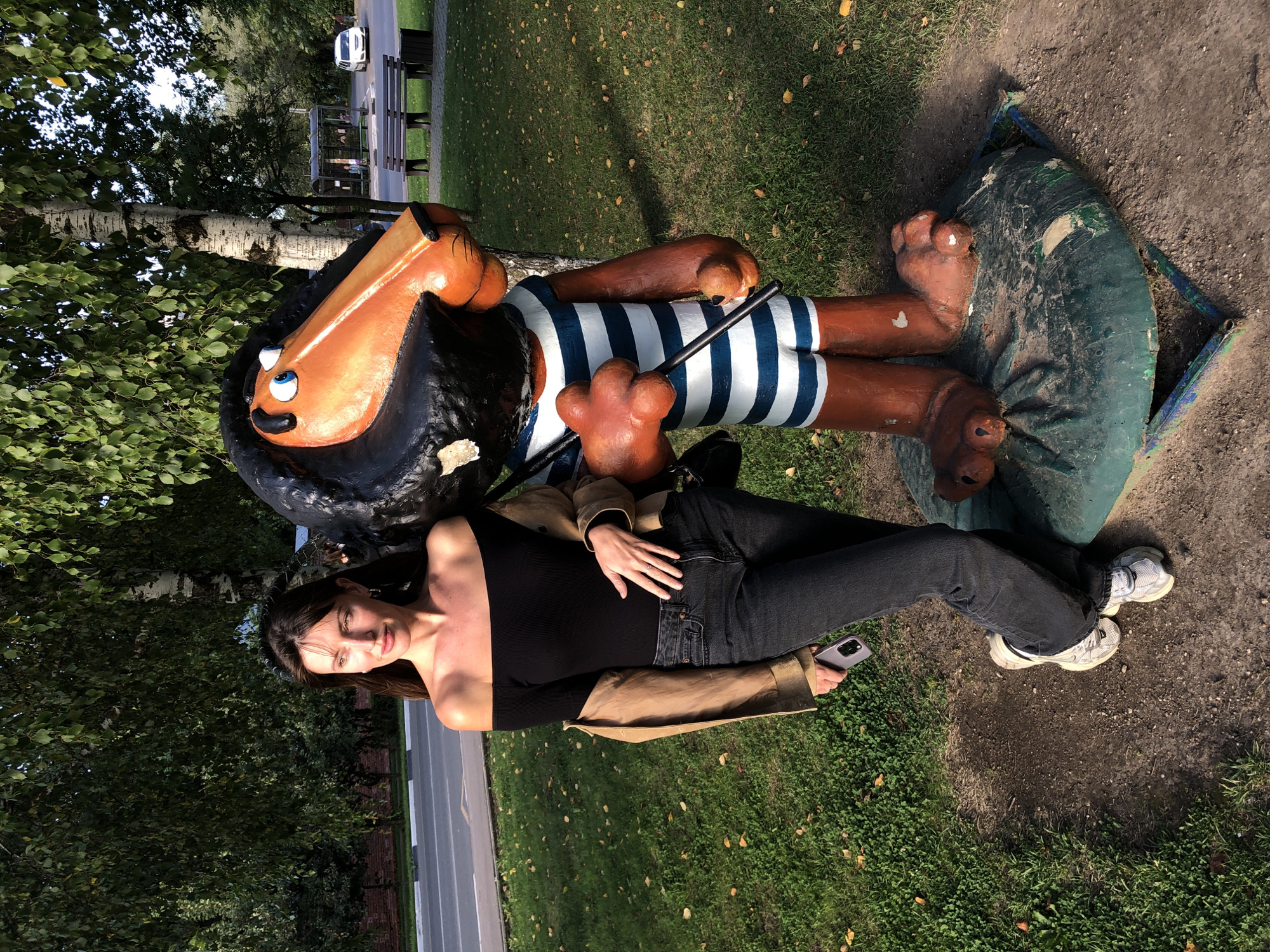 In Sergiyev Posad, Russia, on a sunny day, a young woman with long brown hair stands next to a large, colorful statue of a cartoon lion. The woman is wearing an off-the-shoulder black top, dark jeans, a tan jacket draped over her shoulders, and white sneakers. She is holding a smartphone in her left hand and has her right hand on her hip. The lion statue is painted with a black mane, an orange face, and a blue and white striped sailor suit. It is holding a black stick. The two figures are situated on a grassy area with fallen leaves scattered around, beside a paved road. In the background, there are trees, park benches, a bus stop, and a white car. The lighting suggests it is late afternoon.