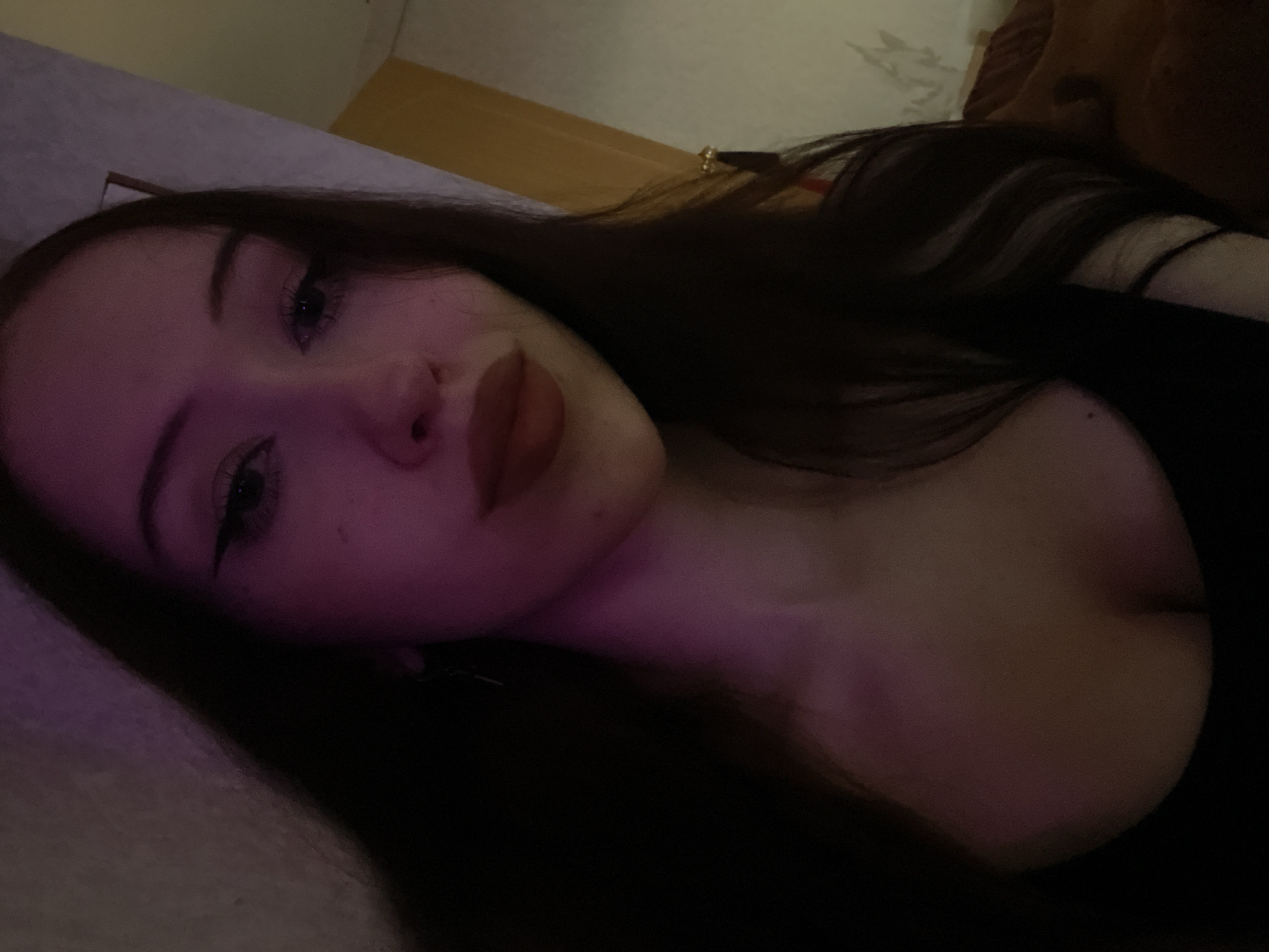 The image is a close-up, low-light selfie of a young woman. She has long, dark hair, dark eyeliner, and is wearing dark lipstick. A faint purple light illuminates her face from the left side, casting shadows. Her eyes are open, and she is looking towards the camera. Her décolletage is visible. The background is indistinct, with hints of a wall and a doorway. There is no indication of the specific location within Votkinsk, Russia.