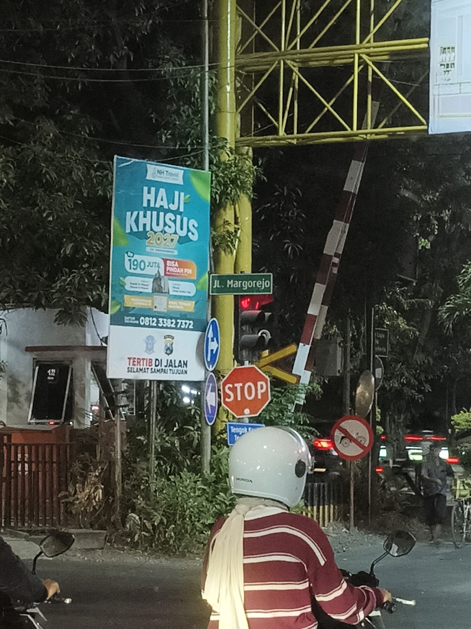 The image depicts a street scene at night in Surabaya, Indonesia, likely near a railway crossing. A yellow metal structure, possibly part of a railway signal or overhead gantry, is visible. A red and white striped boom gate hangs downwards, indicating a train is expected or passing.
A large blue banner advertises "HAJI KHUSUS 2027" and "NH Travel," offering information about a special Hajj package and contact details. Below this banner, another sign reads "TERITIB DI JALAN SELAMAT SAMPAI TUJUAN" (Orderly on the Road, Arrive Safely).
Several traffic signs are present: a green street sign labeled "Jl. Margorejo," a red octagonal "STOP" sign, and a circular sign with a diagonal bar over a bicycle symbol, indicating "No Cycling." A traffic light shows red and yellow lights.
In the foreground, a person wearing a white helmet and a maroon and white striped shirt is seated on a motorcycle, facing away from the camera. The person is wearing a light-colored scarf draped over their shoulders. Another person is partially visible on a motorcycle to the left. In the background, several vehicles with their headlights and taillights illuminated are stopped, suggesting the traffic light or railway crossing is active. Lush green foliage surrounds the area, and the lighting suggests it is evening or nighttime.