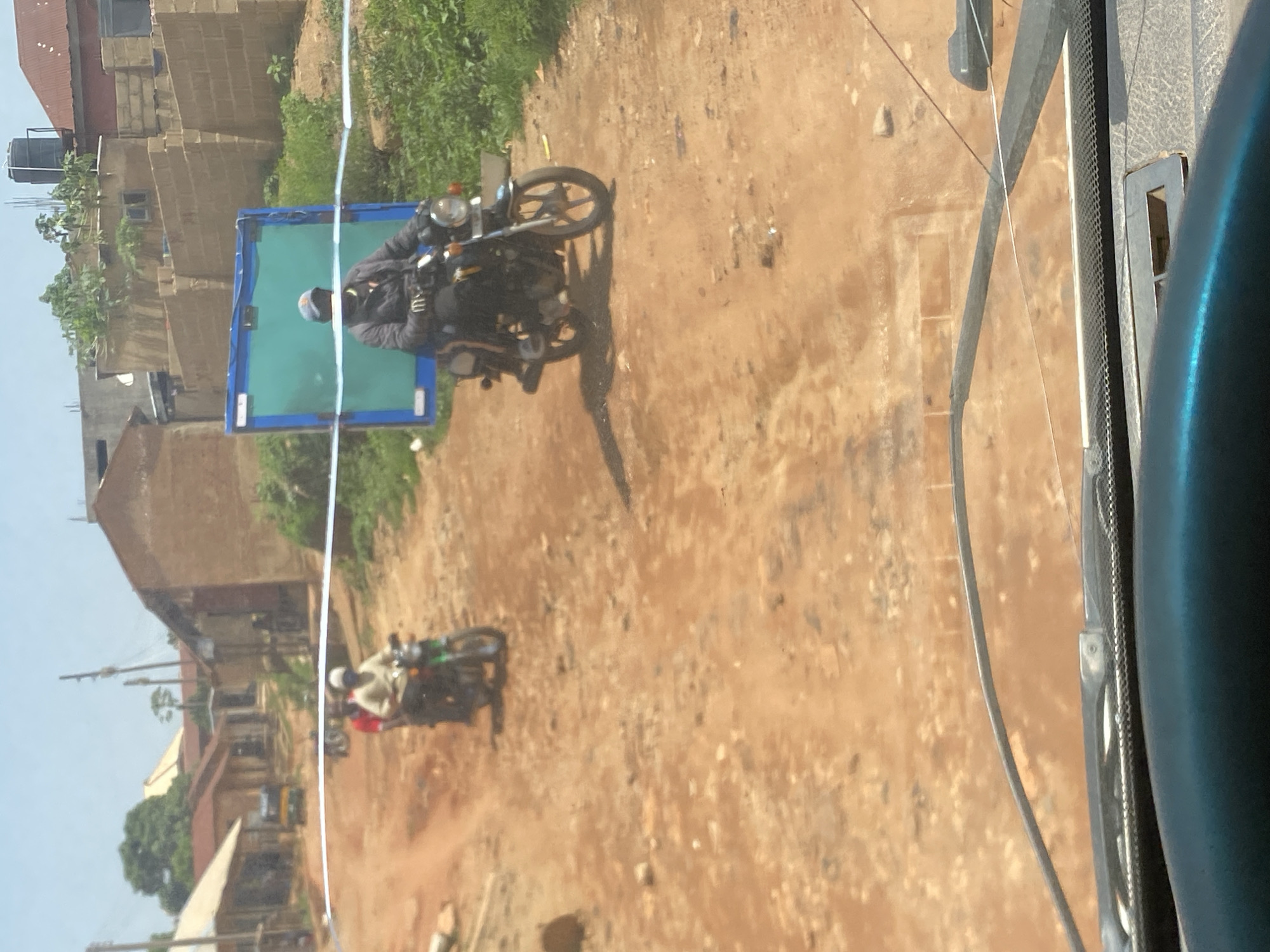 The image depicts a street scene in Jos, Nigeria, characterized by a dirt road and buildings constructed from brick and corrugated iron roofing. The perspective appears to be from inside a vehicle, possibly a car, looking through a windshield that reflects some of the surrounding environment.
The primary subjects are two individuals riding motorcycles. The first motorcycle, closer to the foreground, has a rider wearing a dark jacket and a cap. This motorcycle is positioned near a large, blue rectangular object, possibly a billboard or a covered structure. The second motorcycle is further down the road, with a rider wearing a lighter colored shirt and a red garment.
The environment suggests a warm, sunny day, indicated by the clear sky and the lighting on the road and buildings. The road itself is unpaved and dusty, typical of many areas in Jos. The buildings vary in architectural style, with some appearing older and more weathered, while others seem more modern. Lush green vegetation is visible in some areas, contrasting with the earthy tones of the road and buildings.
The activity is the movement of the motorcycles along the road, suggesting daily transport or travel within the city. There are no discernible emotions evident from the subjects. The scene captures a glimpse of everyday life in Jos, highlighting the local infrastructure and the modes of transportation. No visible text is present in the image.