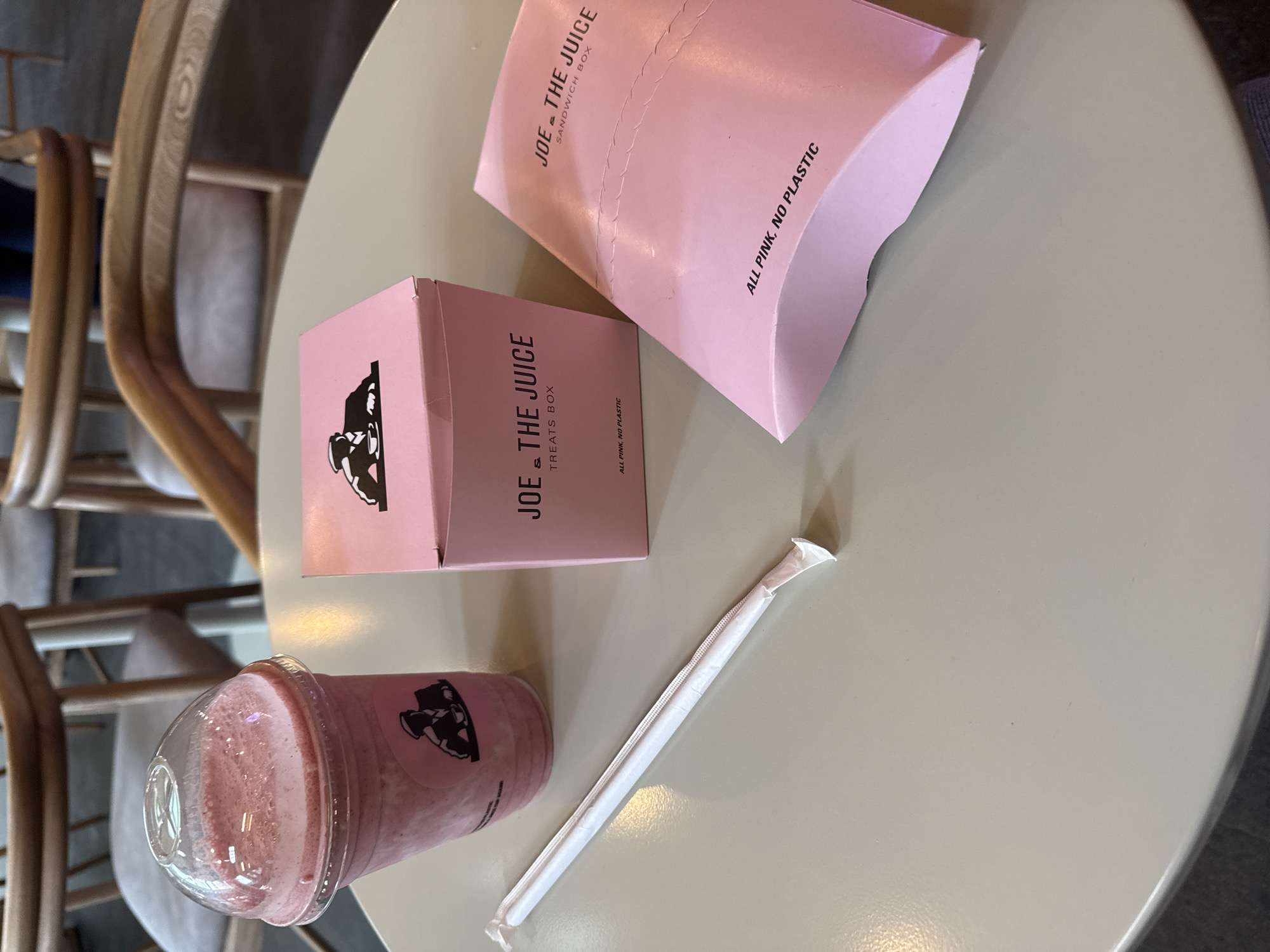 Here is a detailed description of the image:
The image captures a table setting, likely in a café or restaurant in Mexico City, Mexico. The table is round and light gray, with a few items placed on its surface.
There is a pink smoothie in a clear plastic cup with a black logo that resembles a man's face. It appears to be a beverage from "Joe & The Juice," indicated by the branding on a pink box, located closer to the top left corner of the frame. The pink box is labeled as "TREATS BOX." A second pink box, likely a sandwich box, is placed diagonally above the treats box. There is a wrapped paper straw placed near the smoothie.
Behind the table, a portion of a wooden chair is visible, with light gray upholstery. The scene is well-lit, and the focus is sharp on the items on the table. The overall setting suggests a casual dining or refreshment area, possibly within a commercial space in Mexico City.