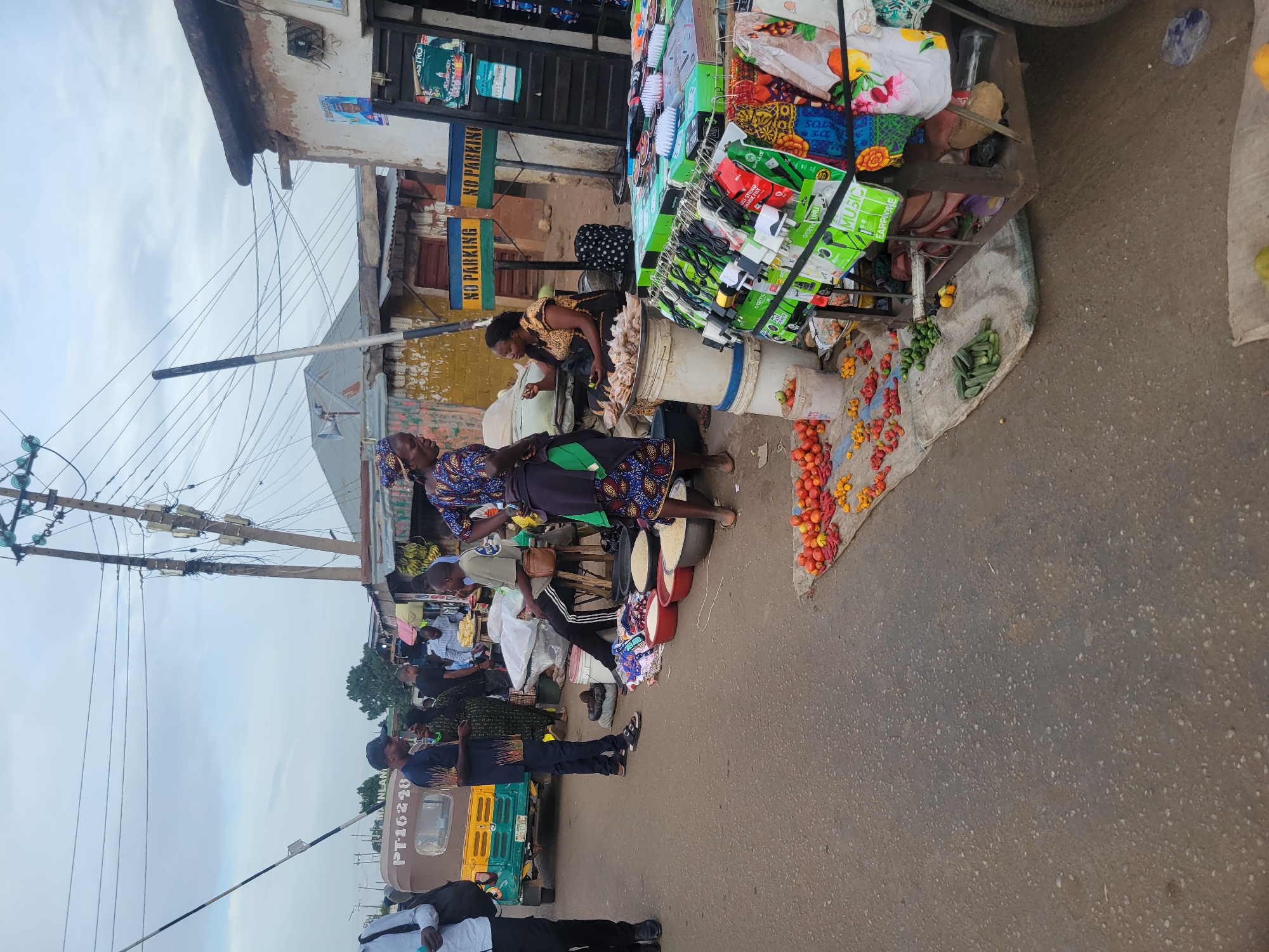 The image depicts a bustling street market scene in Jos, Nigeria, under an overcast sky. The setting is a paved road flanked by older buildings and a utility pole dense with overhead electrical wires.
In the foreground and midground, several people, predominantly women in patterned attire, are engaged in market activities. A central market stall features a display of various packaged goods, including electronics accessories like "oralmo MUSIC EARPHONE" products and chargers. On the ground adjacent to this stall, fresh produce such as red and yellow tomatoes, peppers, and green vegetables (possibly okra) are laid out on a burlap sack. Buckets containing other produce or goods are also visible. One woman is actively attending to this stall, while another stands nearby.
Further down the street, more individuals can be seen, some seated and others standing, indicating an active communal space. A yellow and green motorized tricycle, a common form of transport locally known as a Keke Napep, with the license plate "PT-16218," is visible on the road.
Notable details include "NO PARKING" signs affixed to a building, as well as partial advertisements for "ASTRO" and product brands like "UMU" and "DE TOS" on the market goods. The overall scene captures the vibrant and everyday commerce of a West African street market during daytime.