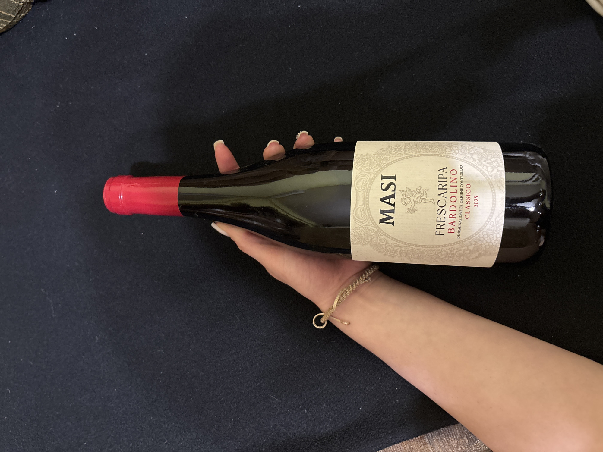 A photograph captures a wine bottle being held by a person's hand against a dark background. The bottle has a red foil cap and a label that reads "MASI FRESCARIPA BARDOLINO CLASSICO 2025". The person has light skin tone and is wearing a bracelet with a loop. The background appears to be a dark-colored, possibly fabric, surface. The location in the city is Unknown, Unknown.