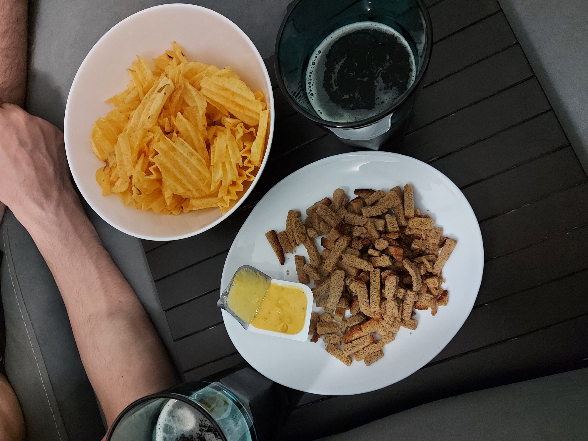 A high-angle, close-up shot depicts a casual snack arrangement. A white bowl contains ruffled potato chips. Next to it is a glass of dark beer with visible foam. A white plate holds rye croutons and a small, opened container of yellow sauce, likely mustard or cheese-based. Part of a person's arm is visible to the left, holding another glass of dark beer. The setting appears to be indoors on a dark wooden-like surface, possibly a small table or tray. The photo was likely taken in Novosibirsk, Russia.