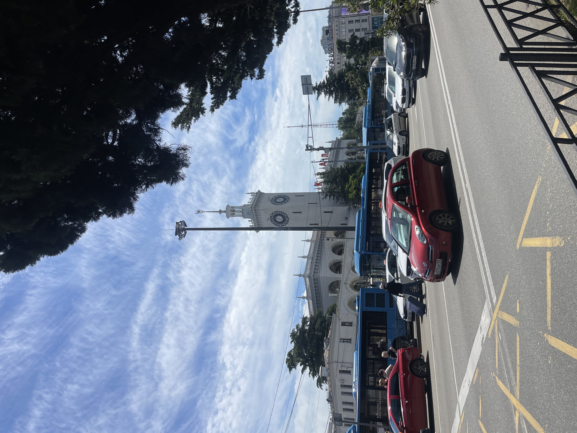 The image shows a street scene in Sochi, Russia. The distinctive clock tower of the Sochi Marine Terminal is visible in the background, set against a blue sky with cirrus clouds. Along the street, several blue buses are parked, and people are standing near them. A red car is parked alongside the buses. Additional parked cars are seen further down the street. The roadway has yellow markings. A tree branch is visible in the upper left corner of the image. A fence is on the right side of the roadway. Overhead power lines are also visible in the sky.