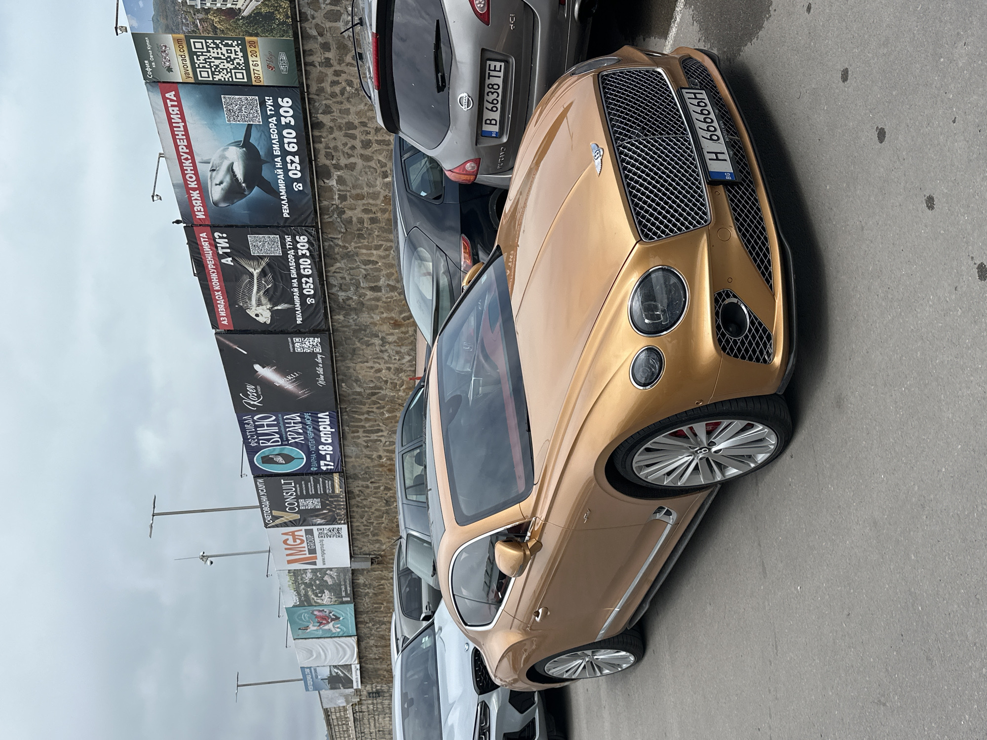 A gold Bentley Continental GT is parked on an asphalt surface, its front facing slightly left. Its Bulgarian license plate reads "H 66666H". Behind it, a silver Nissan Murano with the Bulgarian license plate "B 6638 TE" is visible, along with other partially seen parked vehicles. The scene is set on an overcast day.
To the left of the cars, a tall stone wall is covered with numerous vertical billboards displaying various advertisements. These include promotions for real estate ("yavorad.com," "София кв. Овча Купел"), competitive services featuring images of a shark and a fish skeleton with slogans like "ИЗЯЖ КОНКУРЕНЦИЯТА" (Eat the competition) and "АЗ ИЗЯДОХ КОНКУРЕНЦИЯТА А ТИ?" (I ate the competition, and you?). Contact numbers for billboard advertising ("052 610 306," "0877 61 20 20") are prominently displayed. Other advertisements feature "СЧЕТОВОДНИ УСЛУГИ" (Accounting Services) by "CONSULT GROUP" and "MGA GROUP" (www.mgagroup.bg). A "ФЕСТИВАЛ ВИНО ХРАНА" (Wine Food Festival) is advertised, indicating it will take place at "ВАРНА - ХОТЕЛ ЧЕРНО МОРЕ" on "17-18 април" (April 17-18), featuring "Kosev WINERY" and "AKLI Wine tells a story". The overall location is Varna, Bulgaria, as indicated by the festival location and Bulgarian license plates.
