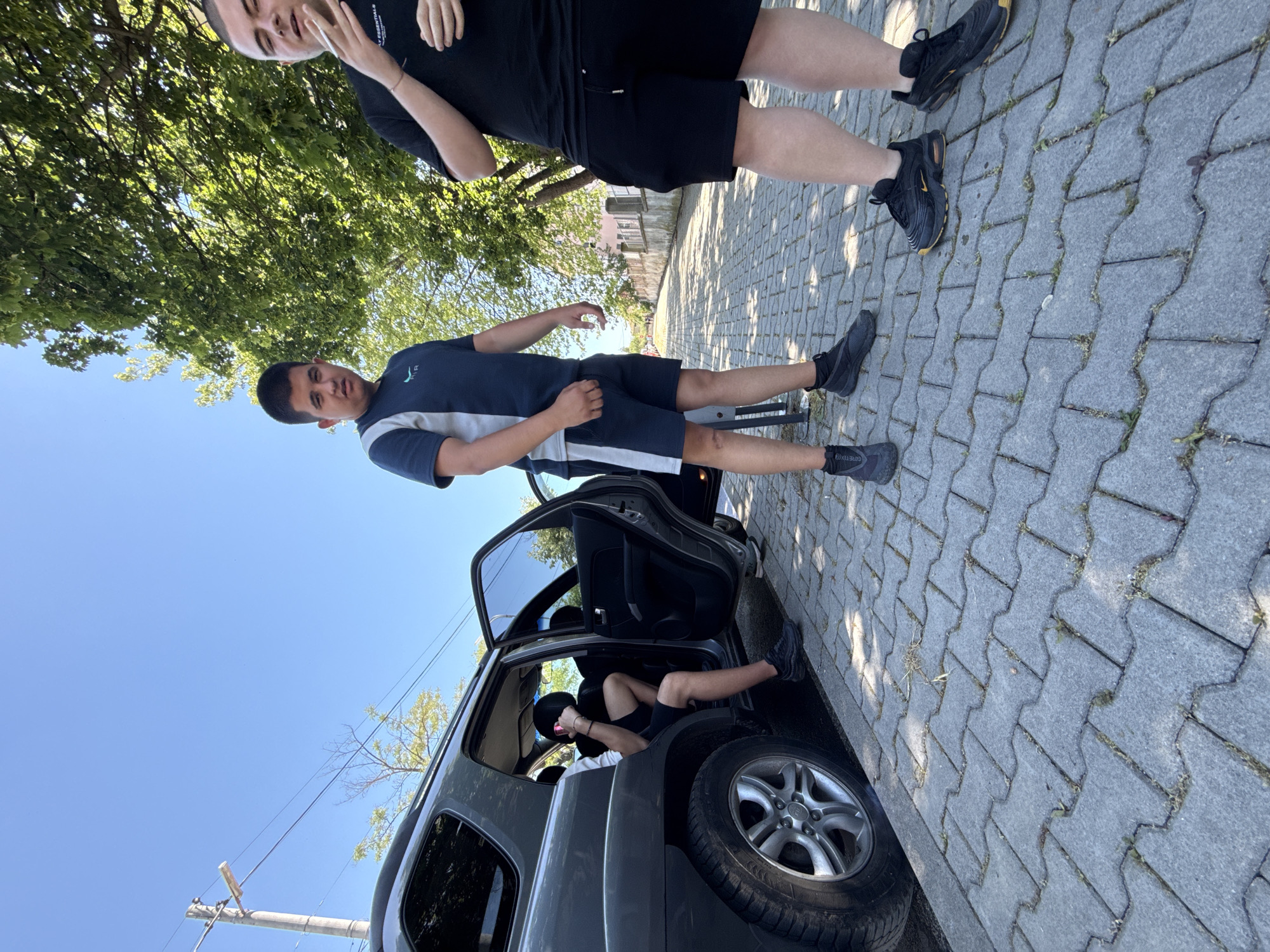On a cobblestone street in Elhovo, Bulgaria, a dark gray SUV is parked with its driver-side door open. A person is inside the car, partially visible in the back seat. Beside the car stands a young man wearing a navy and light gray polo shirt with dark shorts and black sneakers. Another young man, wearing a black shirt, black shorts, and black sneakers, is standing adjacent to him. Trees are visible in the background, and the sky is blue. A utility pole is also present on the left side of the image.