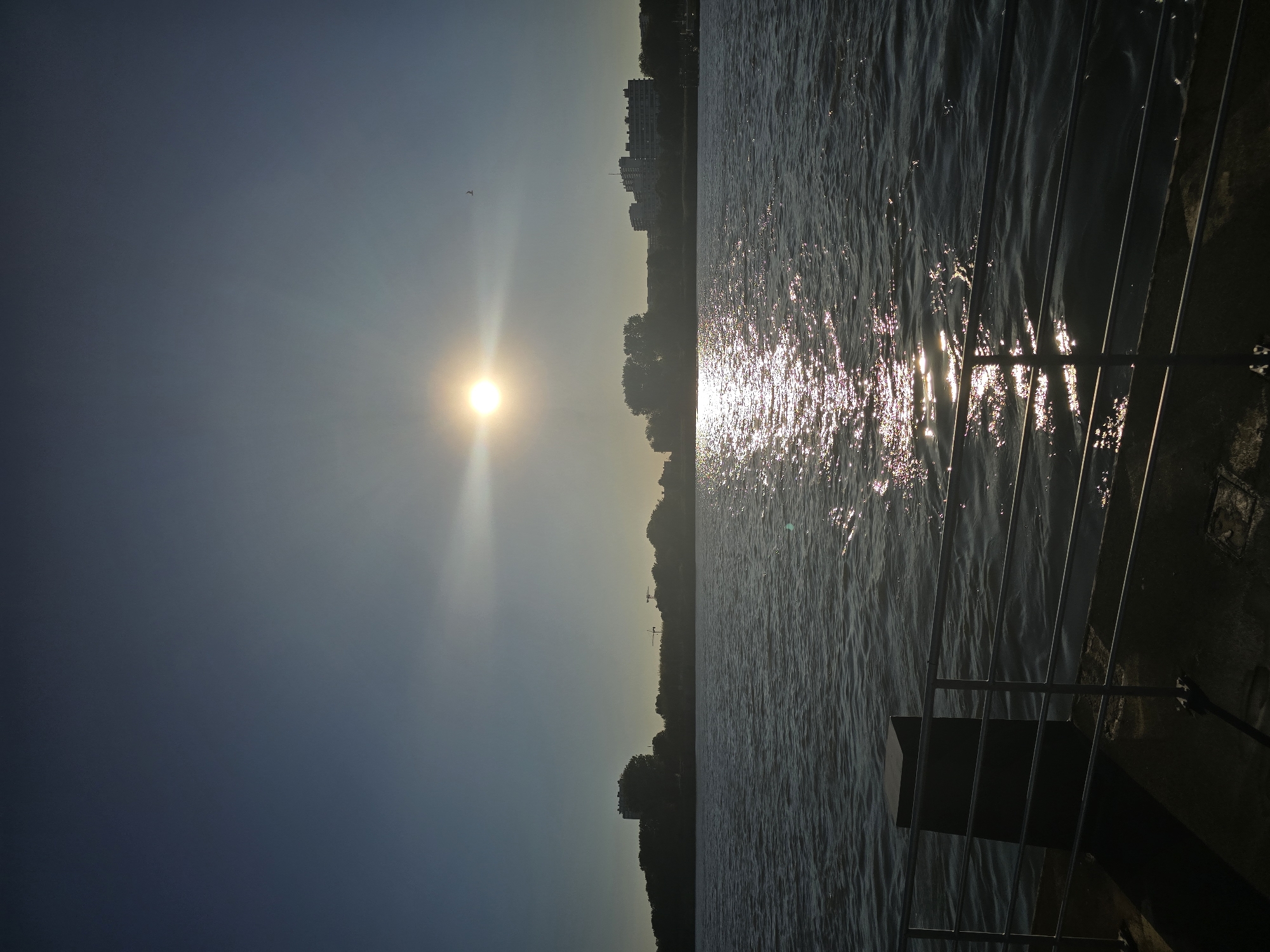 The image depicts a waterfront scene in Antwerp, Belgium, with the sun low in the sky, casting a bright glare on the water. The foreground shows a metal railing and a dark, rectangular bollard on a concrete surface, suggesting a pier or quay. The water is rippled, and a reflection of the sun creates a shimmering path across its surface. In the mid-ground, a silhouetted cityscape with trees and buildings lines the opposite bank of the river. Cranes are visible among the trees, indicating port activity. The sky is clear and blue, with the sun appearing as a bright, almost white orb, surrounded by a lens flare. A single bird is visible in flight high in the sky. The overall impression is one of a calm, early morning or late afternoon view of the port of Antwerp.