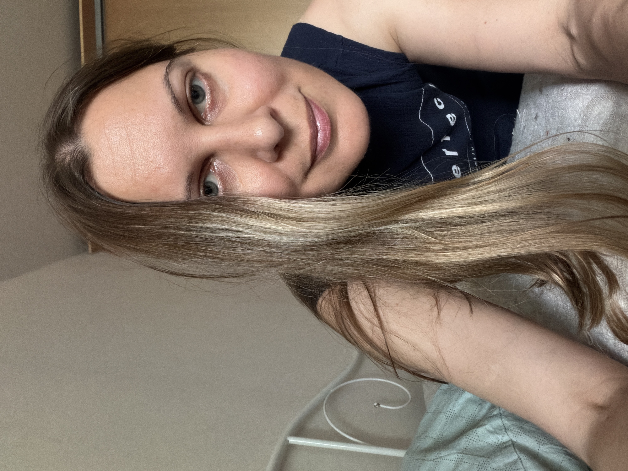 A medium shot shows a woman from the chest up, lying down in bed and looking at the camera. She has long blonde and brown hair that is covering her left shoulder and part of her face. Her eyes are blue and she is smiling. She is wearing a dark blue t-shirt with white lettering. The background shows a wall, a white headboard with a curl on the side, and a light blue pillow. The scene is indoors, likely a bedroom, and it is impossible to determine the specific location within the city of Izhevsk, Russia from the visual information provided.