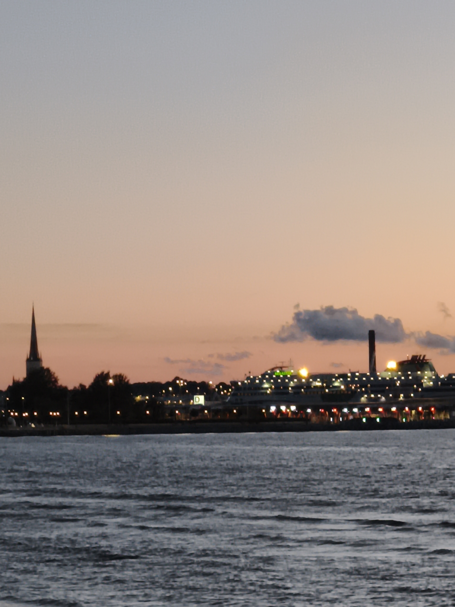 A wide-angle, low-light photograph captures a twilight view of Tallinn, Estonia, from across a body of water. In the foreground, the dark, choppy water reflects the subdued colors of the sky and the distant cityscape.
The middle ground is dominated by the Tallinn skyline, illuminated by numerous lights. On the left, a tall, slender church spire stands out against the dusky sky. Further to the right, a large cruise ship or ferry is docked, its decks and hull adorned with a multitude of bright lights. A smokestack rises from the ship or a nearby industrial building. Behind the ship, more buildings and structures are visible, some also lit up. A white sign with a dark "D" is discernible on one of these buildings. Trees and other vegetation line the waterfront, interspersed with streetlights.
The sky transitions from a pale orange and pink near the horizon to a soft grey at the top of the frame, with a few dark clouds present. The overall impression is of a peaceful evening scene in a coastal city.
