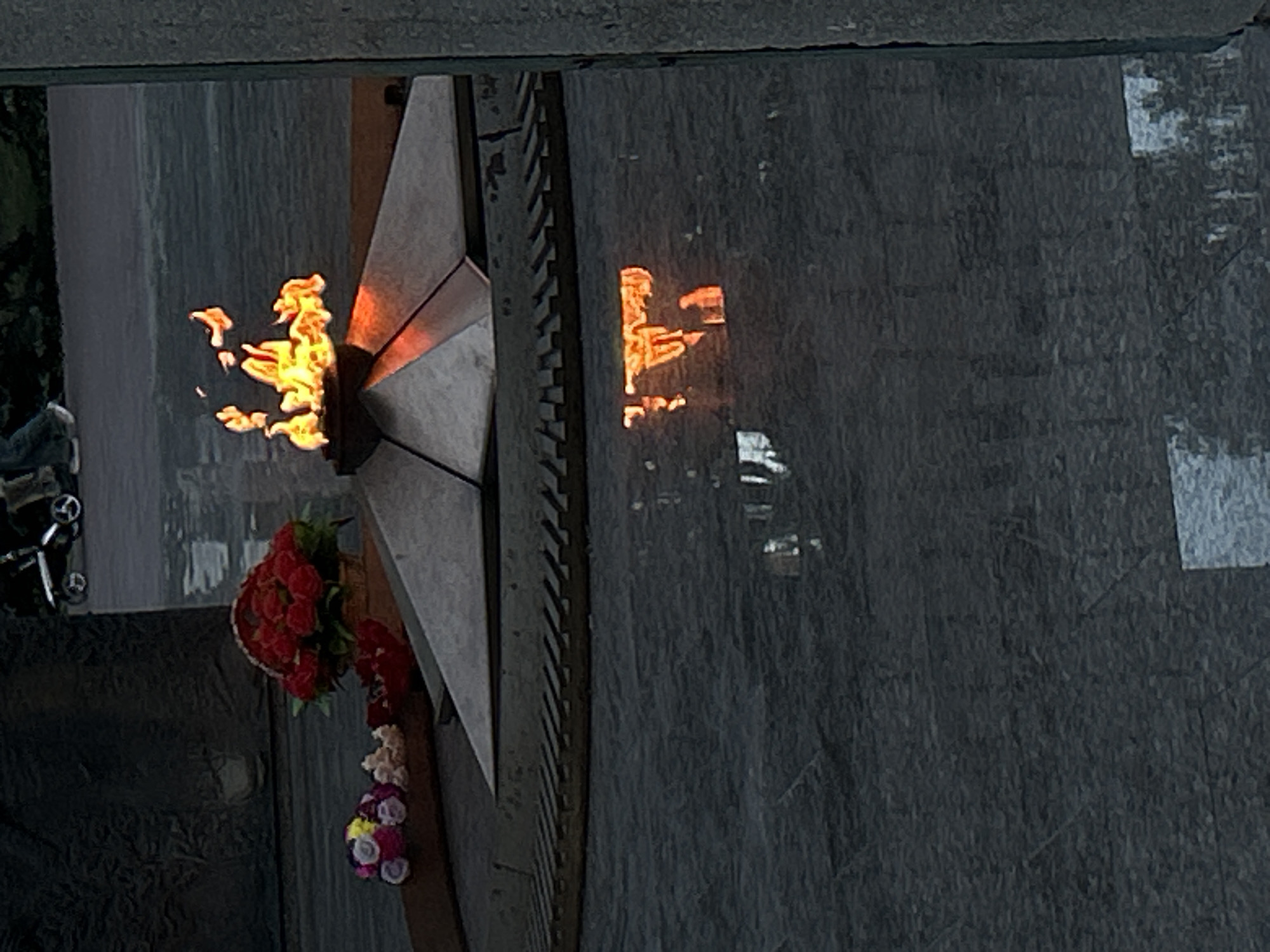 The image depicts the Eternal Flame monument in Tolyatti, Russia. The monument features a five-pointed star with a perpetually burning flame at its center. A flower arrangement is placed near the base of the star. The surface surrounding the monument appears to be stone or concrete with textured markings. A reflection of the flame is visible on the stone surface. Part of a vehicle, possibly a bicycle, is visible on the left side of the image. The overall setting appears to be outdoors.