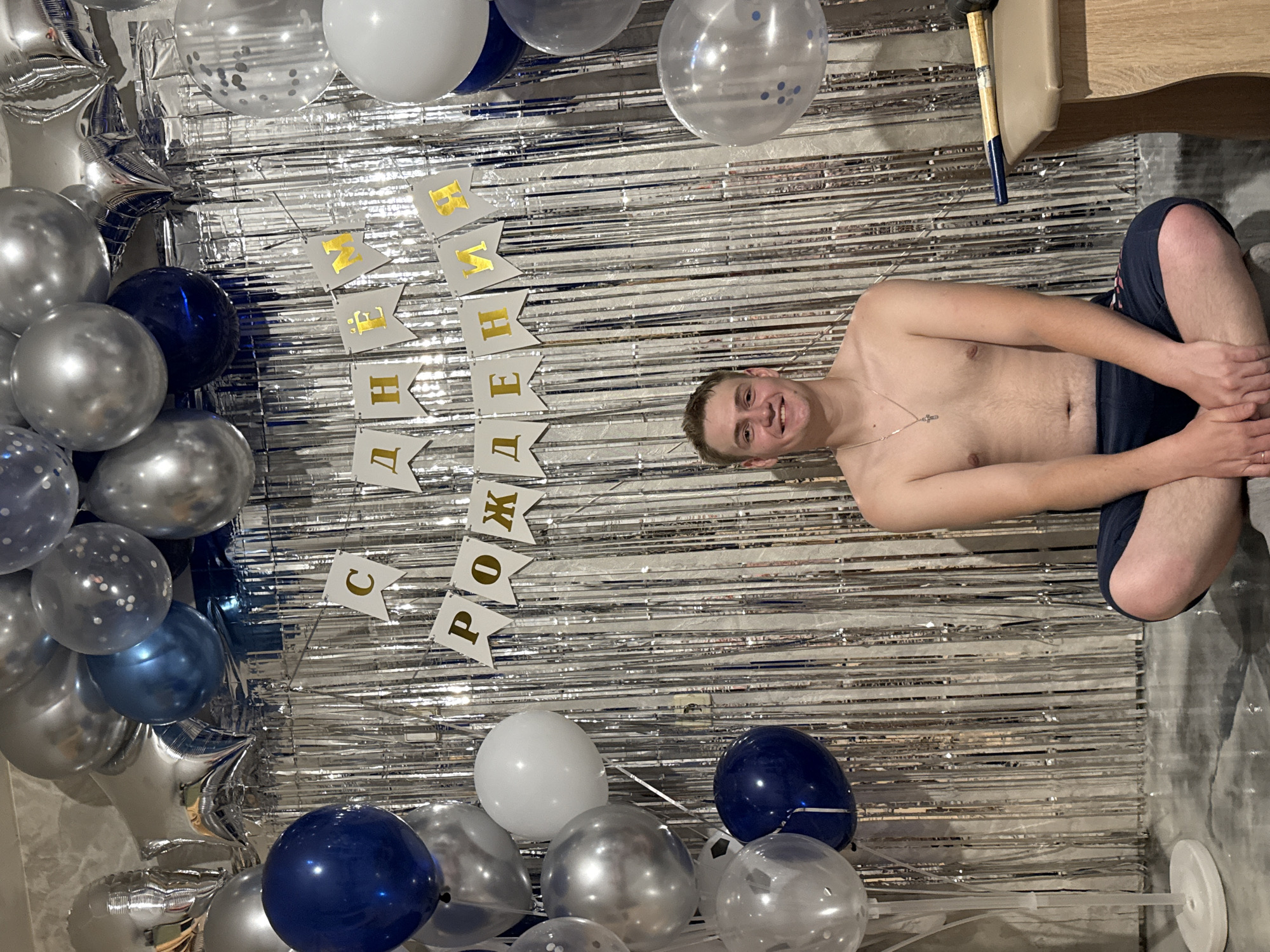 A bare-chested man with a gold cross necklace is sitting cross-legged on the floor with a silver fringe backdrop. The backdrop has white pennants with gold letters that spell out “С Днем Рождения” which translates to “Happy Birthday.” There are silver, white, and blue balloons in bunches to the left and lower center of the image. The room appears to be indoors. The city is Chaikovsky, Russia.