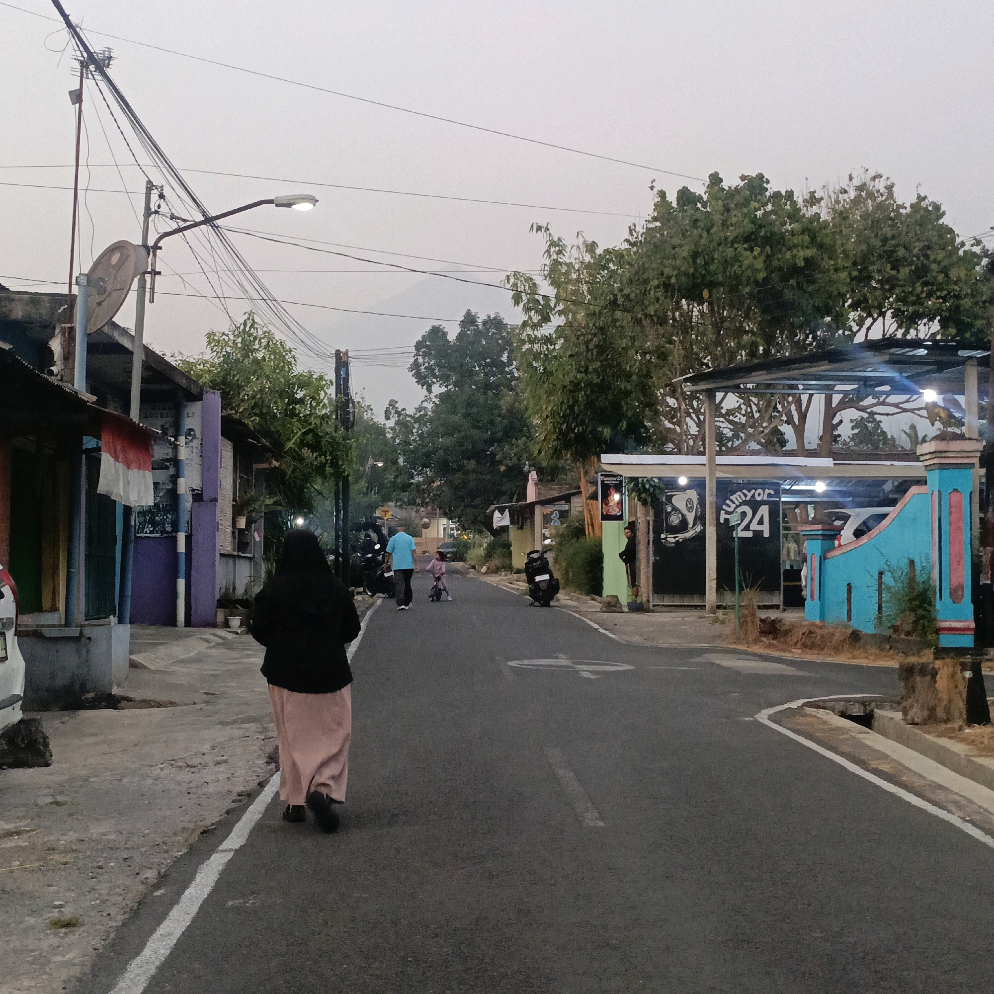 This is a street-level photograph taken in Salatiga, Indonesia, likely at dusk or dawn, with a hazy sky. The scene depicts a residential street with several buildings on the left and right sides. On the left, there are low-rise structures with colorful doors and windows, one displaying an Indonesian flag. A person wearing a black hijab and a long pink skirt walks away from the camera on the left side of the road. Further down the street, a person in a blue shirt walks with a child on a bicycle. Several motorcycles are parked along the roadside. On the right side of the street, there's a building with a blue and pink architectural feature and a sign that reads "Tumyor 24". Trees line the street and are visible in the background, with a faint outline of a mountain in the distance. The road is paved and has white markings. The overall atmosphere is quiet and serene.