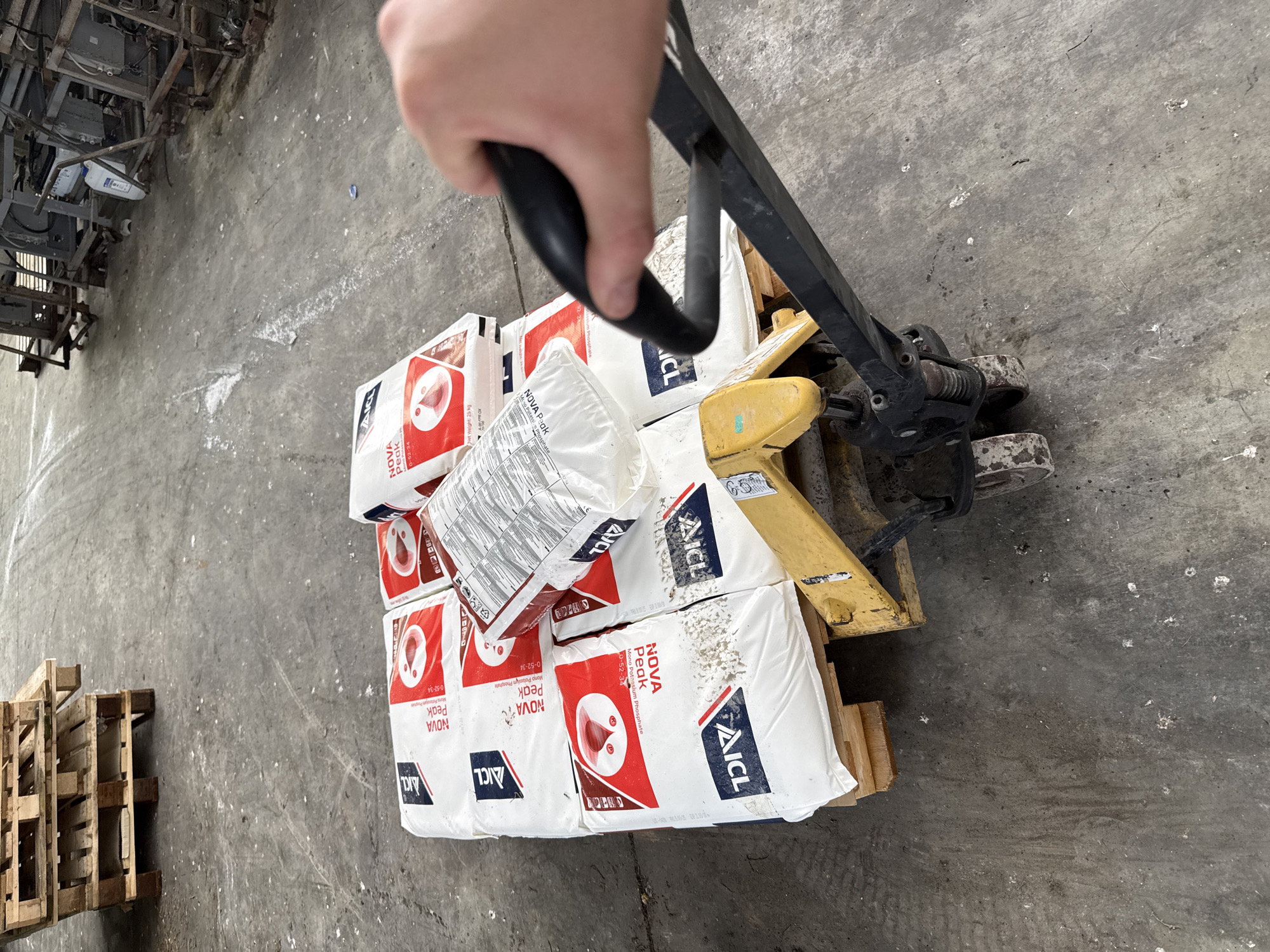 A first-person perspective of a person using a yellow pallet jack to move a pallet loaded with eight bags of Nova Peak fertilizer on a cement floor. The fertilizer bags are white with red and black lettering. Another pallet is visible in the lower-left corner and what appears to be scrap metal on chairs is in the top-left corner. The location is likely a warehouse or storage facility, potentially near Stara Zagora, Bulgaria, given the OCR information on the fertilizer bags.