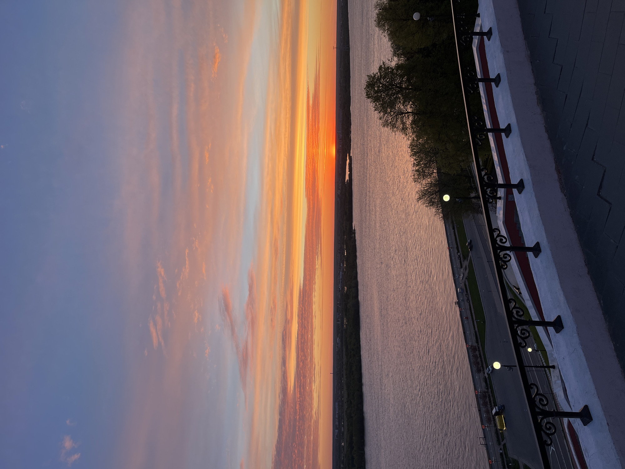 The image shows a sunset over the Volga River in Nizhny Novgorod, Russia. The sky is a mix of blue, orange, and red, with streaks of clouds. The river is reflecting the colors of the sky. Along the bank of the river, there is a paved walkway with ornate black metal railings and lampposts. Trees line the walkway. A road runs parallel to the walkway, with cars driving on it. The perspective is from an elevated position, possibly a building or embankment.