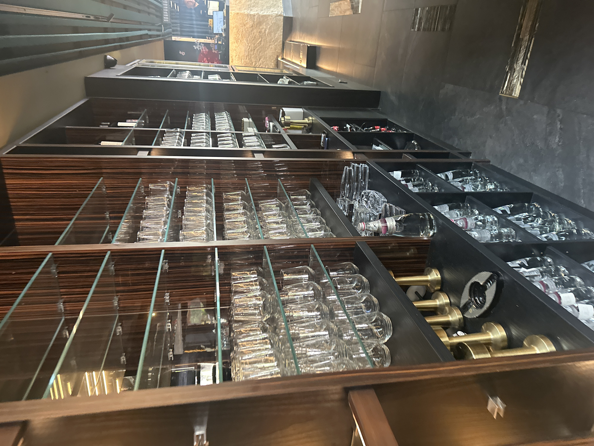 The image shows a wall of shelves in what appears to be a bar or restaurant. The shelves are made of glass and wood, and are stocked with various glasses and bottles. On the left side of the image, there are rows of tumblers. In the center, there are more glasses and some decorative items. On the right, there are bottles of water and what appears to be a back bar area with a person visible behind a counter. The floor is dark and tiled. The lighting suggests an indoor environment, likely during operating hours. The overall impression is one of a well-stocked and organized establishment.