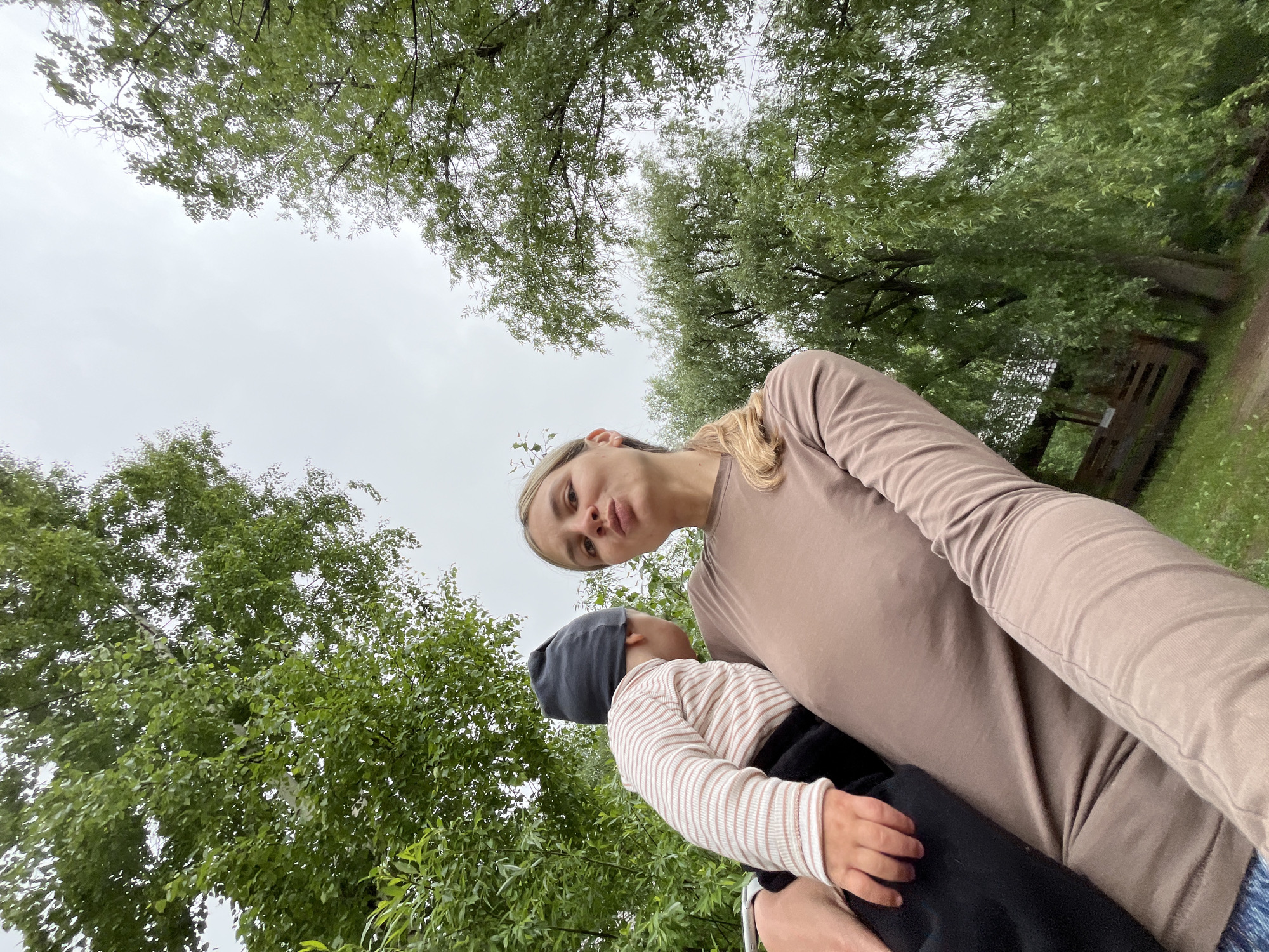 A woman is holding a baby, positioned in front of lush greenery. They are outdoors, with dense tree cover above and around them. The sky is overcast. The location is the city of Chaikovsky, Russia. The woman is wearing a long-sleeved brown shirt. The baby is wearing a striped shirt and a gray hat.