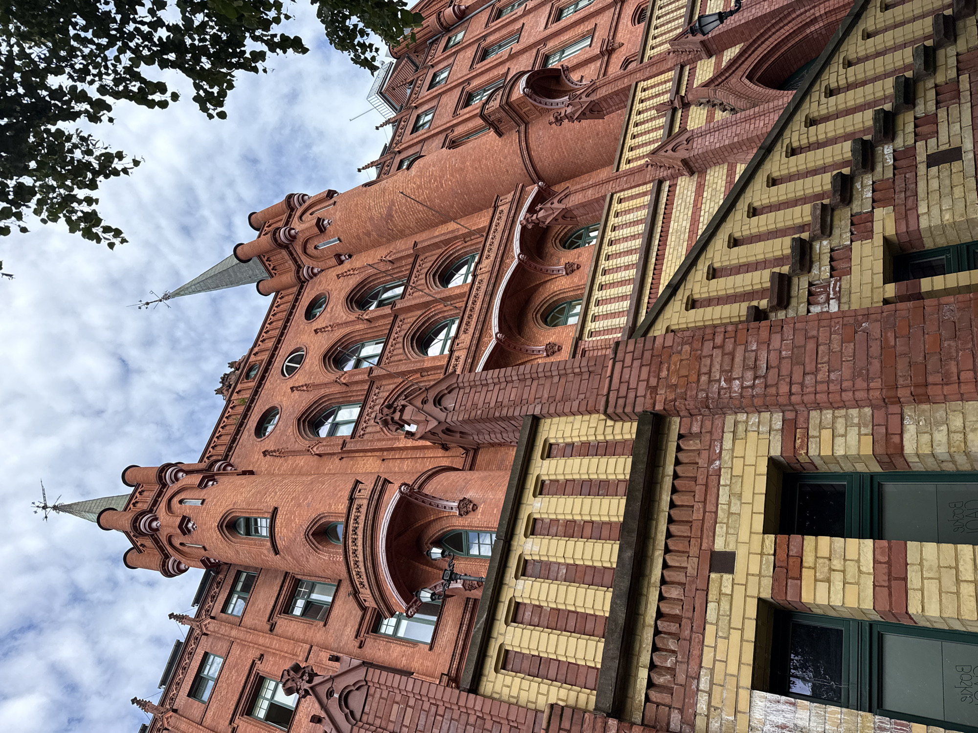 The image captures a low-angle view of a distinctive brick building in Szczecin, Poland. The architecture is characterized by its red brick construction, ornate Gothic-inspired detailing, and prominent spires. The building features arched windows with green frames, decorative balconies, and a staircase with yellow and red brick accents. In the foreground, there are two windows with what appears to be reversed text on them. The sky above is cloudy with patches of blue. A portion of a tree with dark green leaves is visible in the upper right corner. The overall impression is one of historical architecture set against a slightly overcast sky.