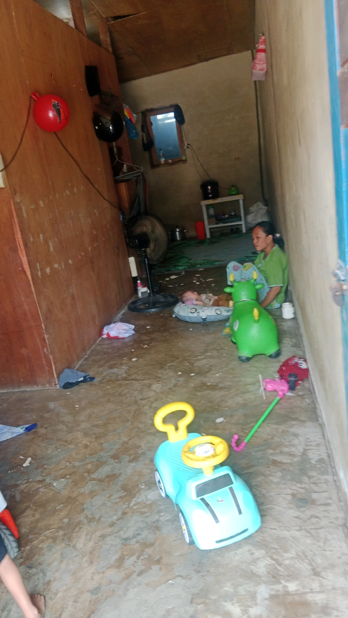 The image depicts an interior scene with a concrete floor and rough walls, suggesting a humble dwelling. A woman in a green shirt sits on the floor, looking towards a baby lying on a patterned mat. Nearby, a green toy cow and a blue toy car are present. A fan is visible in the background. The overall impression is of a domestic space, possibly in a rural or less developed area. The mention of Muda Setia, Indonesia, places the scene in a specific geographic context.