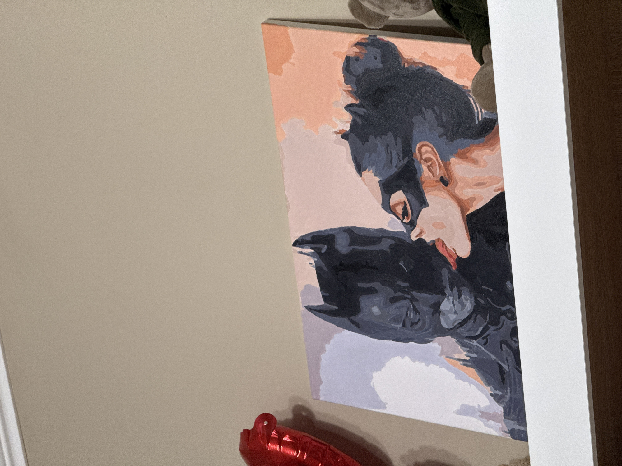 The image shows a paint-by-numbers canvas depicting Catwoman kissing Batman. The canvas is mounted on a wall, likely indoors, and a portion of a red balloon is visible below it. The surrounding wall is a neutral beige. Part of a white shelf or frame is on the right. The location is unknown.