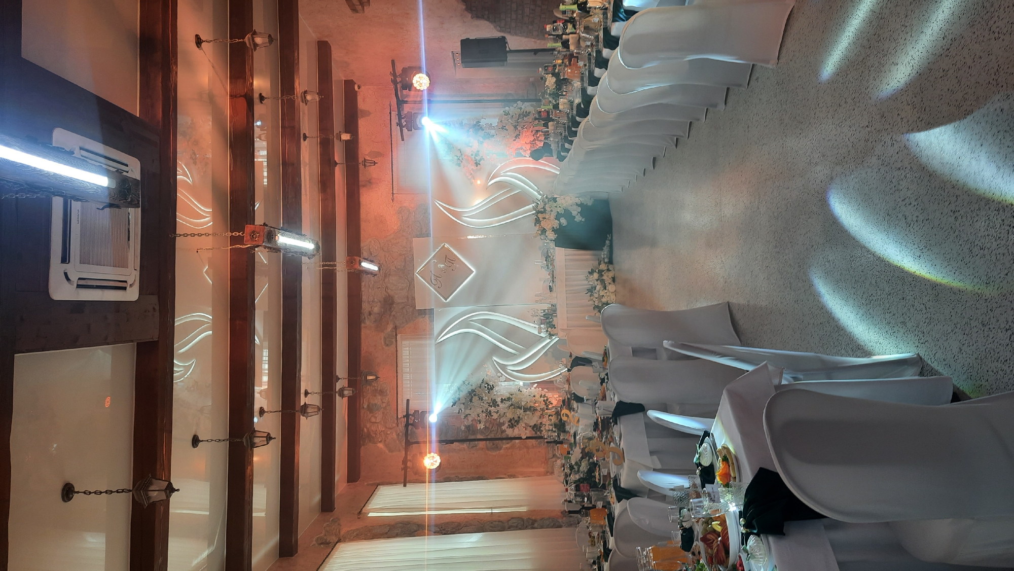 The image depicts the interior of a venue, likely set up for an event such as a wedding reception, located in Жодзішкаўскі сельскі Савет, Belarus. The scene is characterized by a long banquet setup with tables dressed in white tablecloths and chairs covered in white slipcovers. The tables are adorned with floral arrangements, place settings, and drinks. At the far end of the room, a decorated stage area is visible with a backdrop featuring flame-like designs and a diamond-shaped emblem containing initials. Lighting fixtures, including industrial-style lamps and spotlights, are present on the ceiling and stage, casting beams of light and creating illuminated patterns on the floor. The walls appear to be constructed of stone, and the ceiling features exposed wooden beams. The overall atmosphere is one of formal celebration, with emphasis on the decor and lighting to create an elegant ambiance.