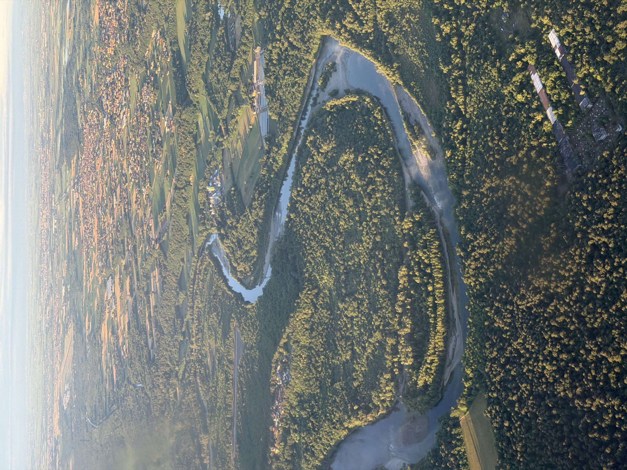 This aerial photograph presents a vast landscape combining natural features with human development, indicative of the region around Pombia, Italy. The primary subject is a wide, light blue river, likely the Ticino, which meanders significantly through the scene, flanked by extensive, dense green forests. Patches of lighter sand or gravel banks are visible along the river's edges.
To the left of the main image, and more distinctly in the first crop, a large residential area spreads across the landscape. Numerous houses with reddish-brown roofs are clustered together, forming a town or village. Adjacent to and interspersed with these settlements are various agricultural fields, appearing as rectangular patches of green and yellow, suggesting cultivated land.
Several man-made structures are visible within the forests. In the second and third crops, long, narrow buildings with light-colored roofs, possibly industrial facilities, warehouses, or large greenhouses, can be seen. One crop also shows a straight, dark line cutting through the landscape, which could be a road or a railway line.
The image suggests a time of day during the golden hour, either late afternoon or early morning, evidenced by the long shadows cast by trees and structures, and the warm, golden light illuminating parts of the terrain. The sky appears clear, though a light atmospheric haze is visible, particularly towards the horizon on the left. No specific human activity is discernible from this high-altitude perspective, nor is any visible text present. The scene conveys a sense of natural beauty and the subtle integration of human habitation within it.