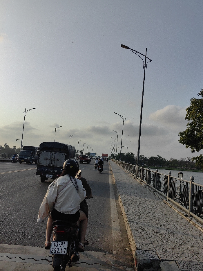 The image depicts a late afternoon or early evening scene on the Phú Xuân Bridge, spanning the Perfume River in or adjacent to Thuận Hóa Ward, Huế City, Thừa Thiên Huế Province, Vietnam.
In the foreground, a dark-colored Honda motorcycle, bearing license plate "43-H1 292.47," is observed from a rear perspective. It carries two occupants. The rear passenger, identified as female, wears a dark helmet, a light-colored, loose-fitting, long-sleeved shirt, dark shorts, and sandals. Her long dark hair is visible beneath the helmet, and she carries a light-colored bag over her left shoulder. The driver, largely obscured by the passenger, wears a dark helmet and dark clothing.
The motorcycle is positioned on a multi-lane asphalt roadway, with traffic flowing in the same direction. Immediately ahead of the motorcycle is a dark green utility truck, license plate "75C-122.70," featuring a canvas-covered cargo area. Further down the bridge, multiple other vehicles, including another similar utility truck, a red pickup truck, and several motorcycles, are visible in motion.
To the right of the roadway, a pedestrian and bicycle path, paved with gray interlocking tiles in a circular pattern, extends along the bridge. This path is bordered by a dark metal bridge railing, which incorporates decorative vertical panels featuring cutout designs. Beyond the railing, the calm surface of the Perfume River is visible, reflecting the pale sky. The distant riverbank is lined with green trees and foliage.
Overhead, a series of tall streetlights with distinct decorative arching fixtures line both sides of the bridge, extending into the distance. The sky is a pale blue-grey with scattered white cumulus clouds, consistent with twilight conditions. Numerous small dark specks, likely birds or insects, are visible against the sky. The streetlights are not illuminated. The overall environment indicates an active urban bridge crossing with moderate traffic.
