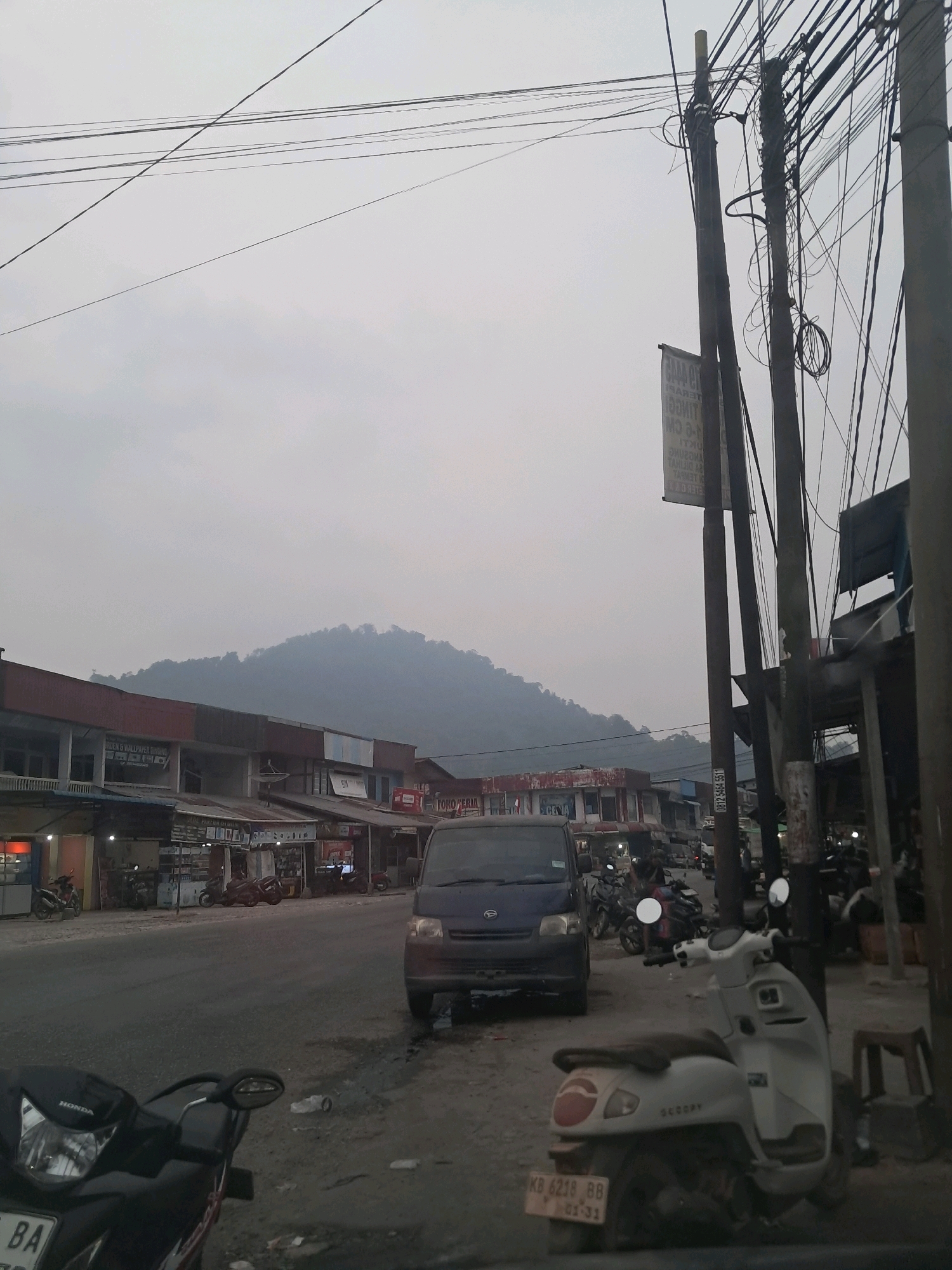The image depicts a street scene in Anjungan Melancar, Indonesia, under an overcast sky. A two-lane asphalt road runs horizontally across the frame, with a mountainous landscape visible in the background. Along the right side of the road are several buildings, including shops with awnings and signs. Power lines crisscross the sky above the street.
In the foreground, a black Honda motorcycle is parked on the right shoulder, facing left. Behind it, a white scooter is also parked, its license plate clearly visible as "KB 6218 BB". A dark blue van is positioned in the middle of the road, facing away from the camera. Several other motorcycles are parked or in motion further down the street. The environment appears to be a commercial area with shops and businesses lining the road. There are no identifiable individuals prominently featured in the scene.