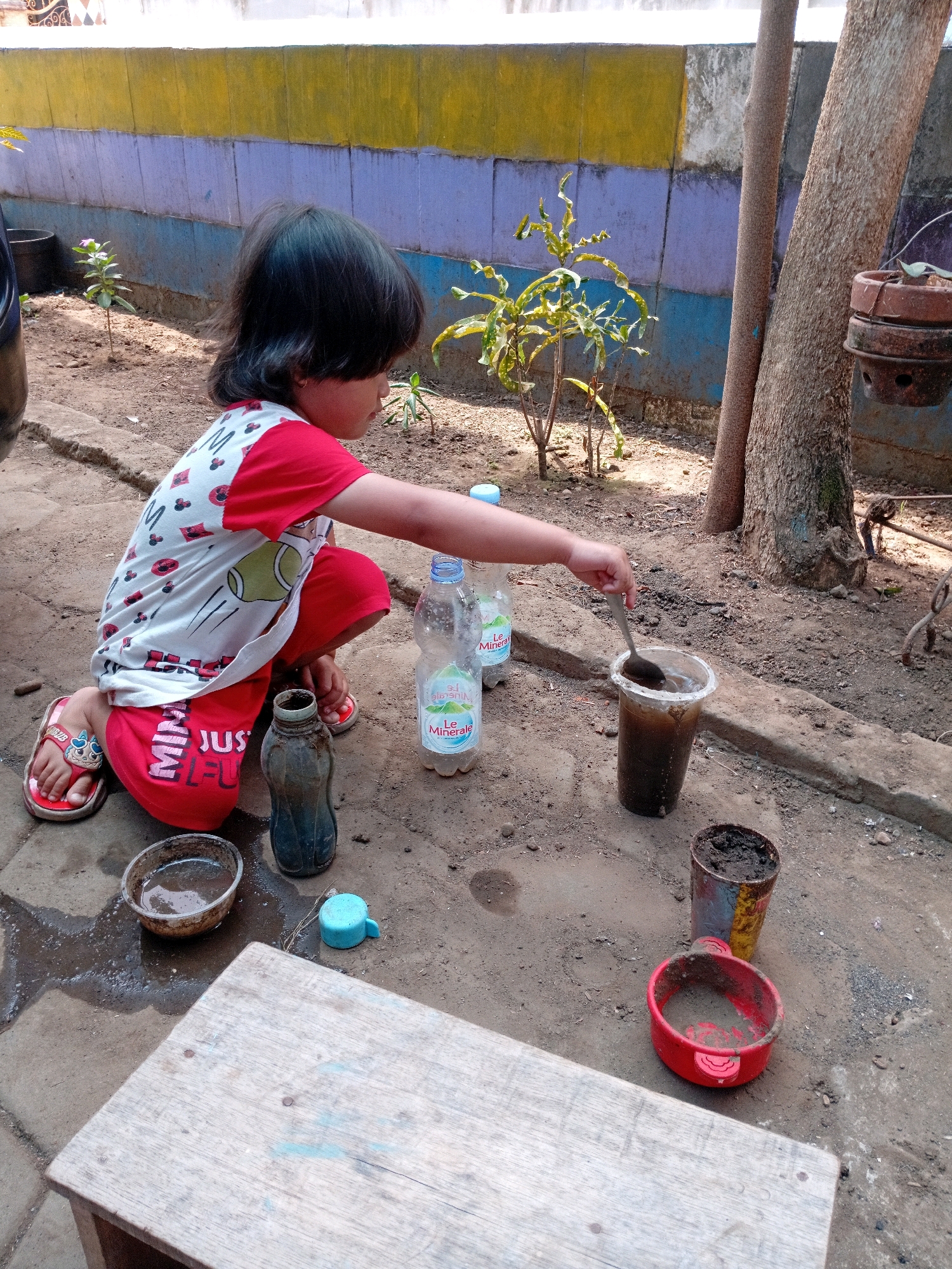 A young child kneels on the dirt ground in Gebang, Indonesia, engaged in an activity with various containers and liquids. The child wears a red and white t-shirt with a pattern and red shorts with "JUST FUN" printed on them. They are wearing red sandals with blue designs. In front of the child is a clear plastic cup filled with murky liquid, and the child is holding a spoon in the cup, appearing to stir the contents. To the left of the cup is a dark glass bottle with dark liquid inside. Further to the left is a small, clear bowl with some liquid. Beside the glass bottle is a small blue plastic object, possibly a lid or a toy. Behind the child, to the right, are two plastic water bottles, one labeled "Le Minerale". In the background, there is a wall with horizontal stripes of yellow, purple, and blue. Trees are visible behind the wall, and a brown hanging pot is attached to one of the trees. The ground is made of dirt and stones, with some scattered leaves. To the lower left, a corner of a wooden bench or table is visible. The overall scene suggests an outdoor, possibly yard or garden setting.