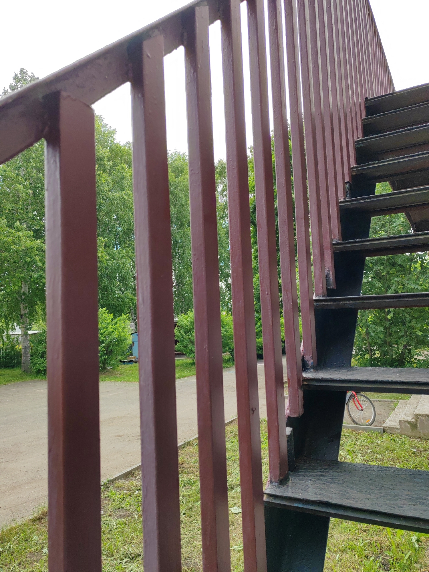 A close-up, slightly upward-angled view captures a dark red-brown metal railing and a black metal staircase in an outdoor setting. The railing, composed of evenly spaced vertical rectangular bars, occupies the left and central parts of the frame, gradually ascending towards the right where it meets the black metal staircase. The stairs are robust, with textured steps.
Through the gaps in the railing and beneath the staircase, a vibrant green landscape is visible. Lush, leafy trees, some with light-colored trunks resembling birches, dominate the background. Below the trees, there's green grass and a light-colored dirt path or road with a concrete curb.
A notable detail is a bicycle wheel, featuring a bright red rim and a white tire, partially visible on the grass directly beneath the lower steps of the staircase. In the deeper background, obscured by foliage, are glimpses of what appears to be a light-colored building and a small blue structure or shed.
The scene is set during the daytime, with bright, diffuse lighting suggesting either an overcast sky or an early morning/late afternoon. There are no people or visible text in the image. The overall impression is that of a functional, possibly industrial, structure situated within a natural or semi-urban green space.