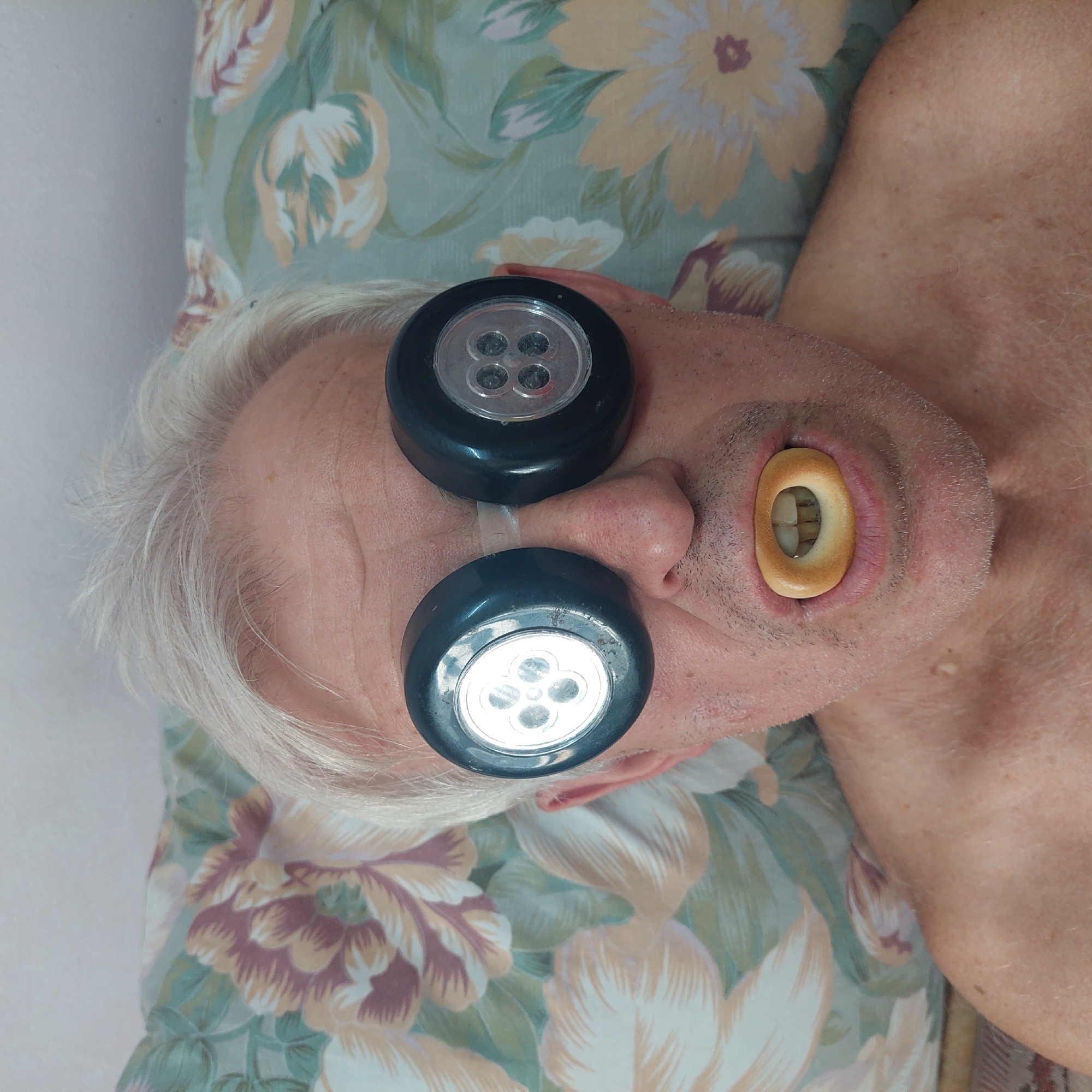 The image shows an overhead, close-up view of an elderly man's face lying on a floral-patterned pillow. He has white hair and is wearing what appear to be two black, circular devices over his eyes, each with a clear plastic front revealing four small, circular lights. Below his nose, his mouth is open, and a bagel is placed inside, encircling his teeth. The lighting is bright, illuminating the man's face and the objects. The floral pattern on the pillow is predominantly in shades of green, yellow, and pink, with large flower motifs. The man's expression is neutral, and the overall scene is unusual and surreal. There is no visible text in the image.
