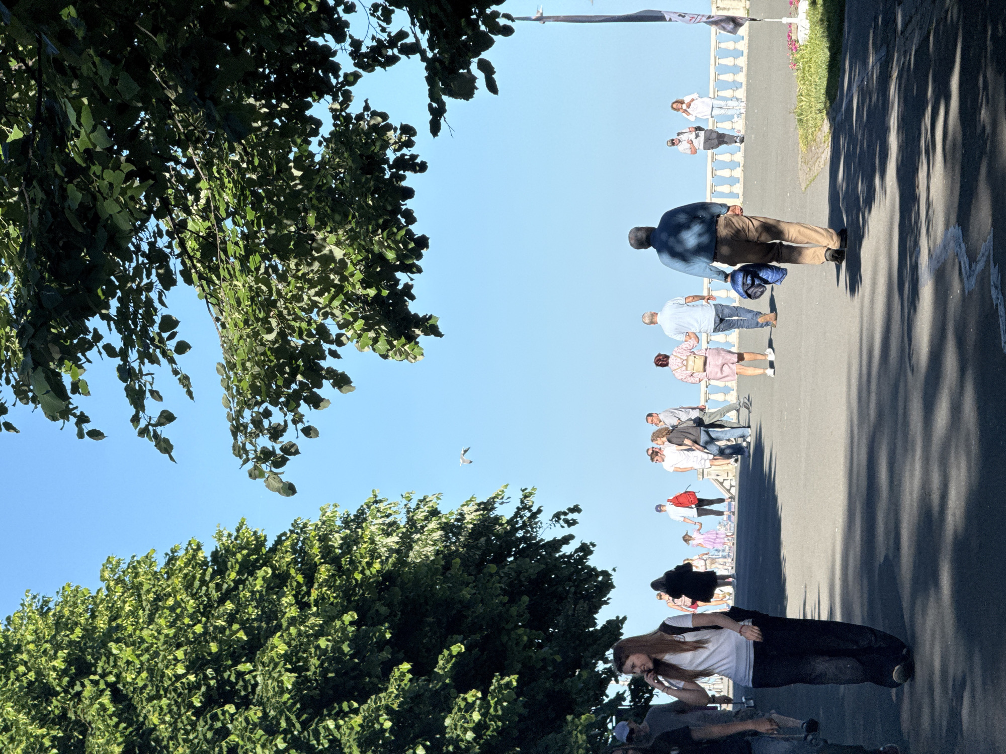 The image depicts a street scene in Burgas, Bulgaria. People are walking along a promenade next to a row of trees under a clear blue sky. The promenade appears to be lined with a low, ornate balustrade or railing. There's a person using a phone. A bird is visible in the sky. The scene is brightly lit, casting shadows on the pavement.