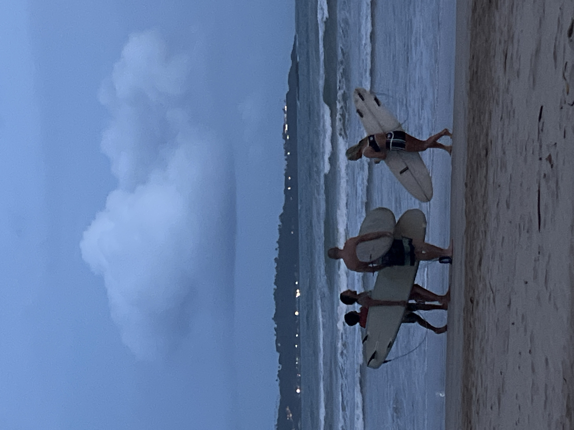 Three individuals are depicted standing in shallow water near a sandy beach, each holding a surfboard. The sky is overcast with large clouds. In the distance, lights are visible, suggesting a coastal city. The location is identified as Peleana, Sri Lanka.