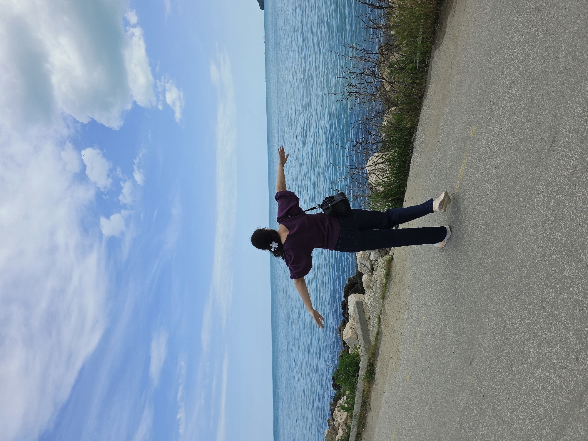 A woman with dark hair, adorned with a white flower accessory, stands on a concrete pathway with a body of water to her left. The woman wears a dark purple shirt, dark pants, and light-colored shoes. Her arms are extended to the sides in a horizontal manner, suggesting a pose or action. A black bag is slung over her shoulder. The water appears calm and blue, stretching towards the horizon under a sky with scattered white clouds. A rocky shoreline with sparse vegetation separates the pathway from the water. The image is taken near the sea in Varna, Bulgaria.
