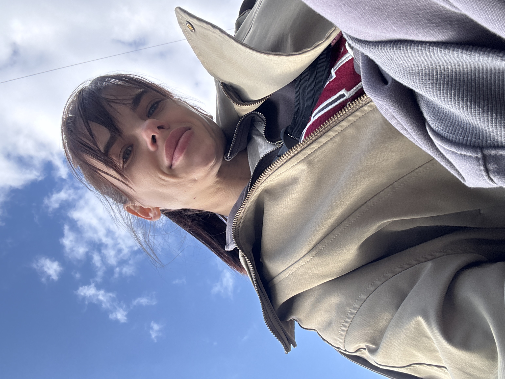 A woman with dark hair and bangs is looking down at the camera, with a sky filled with scattered clouds in the background. She is wearing a light-colored trench coat, partially zipped, and a gray hooded sweatshirt. The angle is a low-angle shot, making her appear to loom over the viewer. No specific location in Rostov-on-Don, Russia, is identifiable from this image.