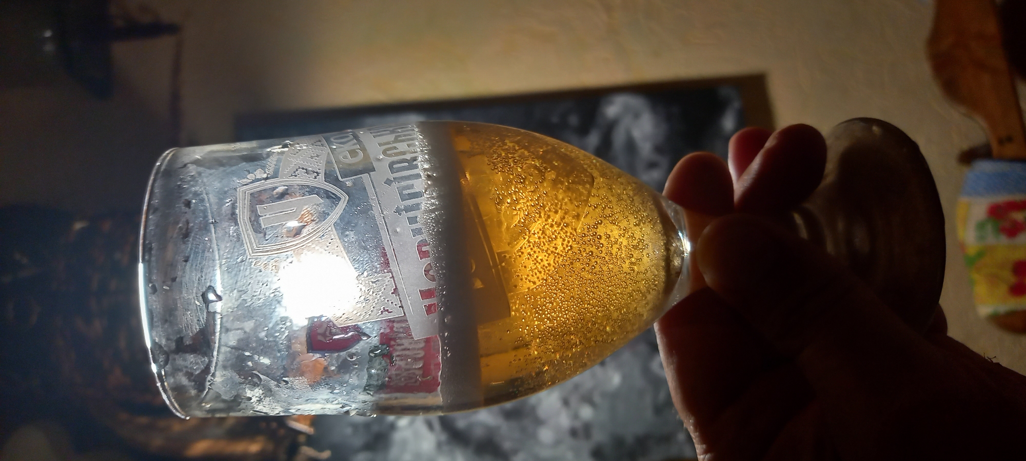 The image captures a close-up, backlit view of a human hand holding a stemmed glass filled with beer, set in an indoor environment in Kryvyi Rih, Ukraine. The glass, positioned centrally, is a pilsner-style glass, exhibiting condensation and water droplets on its exterior, indicating a cold beverage. It contains a clear, golden-yellow liquid with numerous fine bubbles rising from the bottom, consistent with a carbonated alcoholic drink. A thin layer of white foam rests atop the liquid.
Etched or printed on the glass are Cyrillic characters and a shield-shaped logo. The logo features a stylized letter "Ч" or "H" within a shield. Below the logo, partially obscured by condensation and reflection, are the words "Пиво Моравське ЕКО" (Pivo Moravske EKO), indicating a Ukrainian beer brand.
A human hand, primarily silhouetted due to backlighting, grasps the stem of the glass. The hand's fingers are visible, and its skin tone appears dark against the ambient light.
The background elements are largely out of focus. In the upper left, a dark, indistinct object, possibly a decorative figure or sculpture, is visible. To the right, behind the glass, a large, dark, rectangular form, likely a framed picture or poster, displays an abstract, light-colored pattern against its dark surface. The lower background reveals a blurred, light-colored surface, possibly a floor or carpet, and a section of colorful fabric with fruit patterns (cherries, lemons, and blue shapes) to the lower left, possibly a tablecloth or cushion. A dark wooden object, potentially furniture, is vaguely visible to the lower right. The scene is illuminated primarily from behind the glass, creating a strong contrast and highlighting the beer's effervescence.