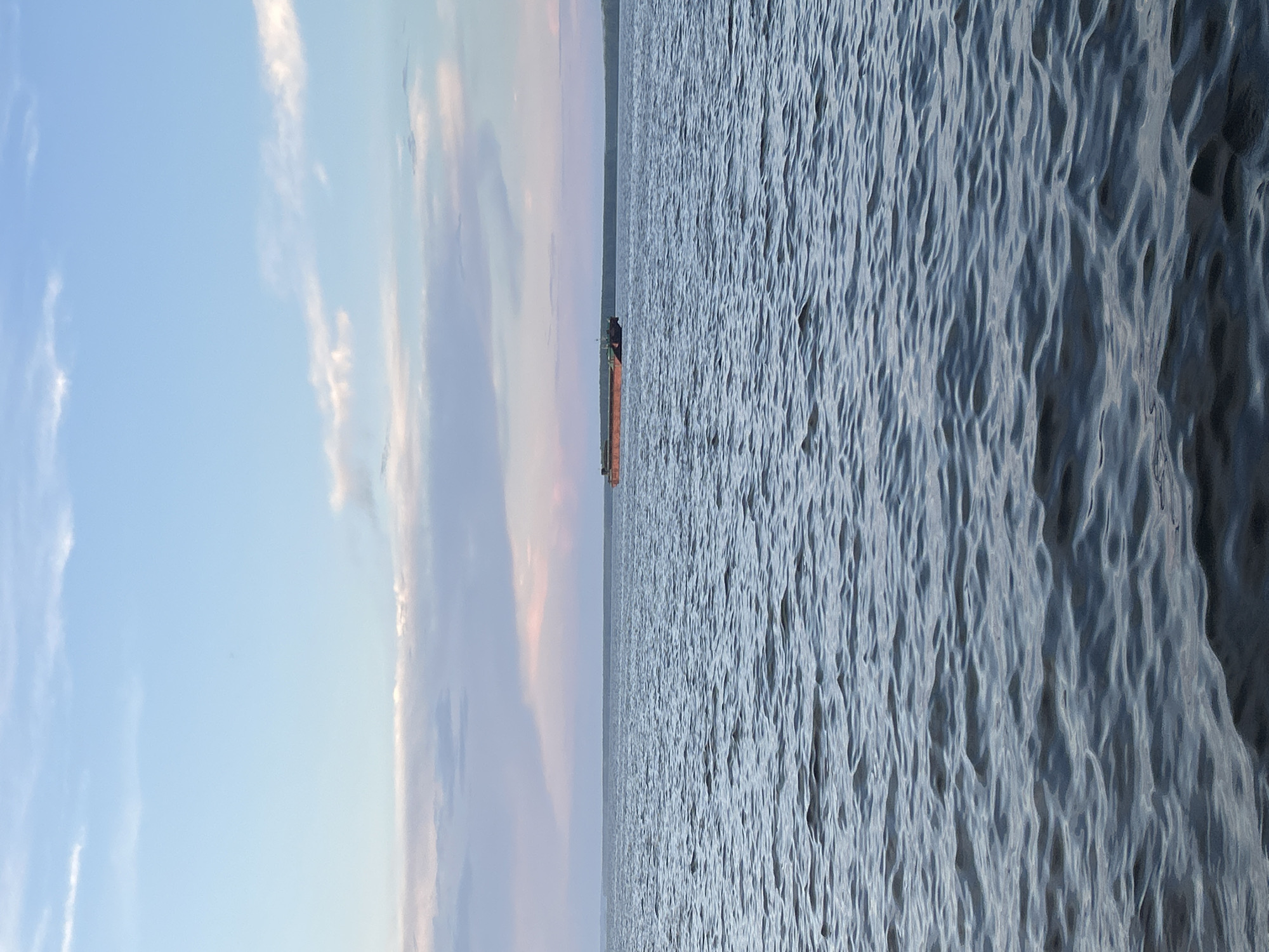 A barge is seen in the distance on a body of water, likely a river, under a sky with scattered clouds tinged with pink and blue hues. The water exhibits moderate ripples, reflecting the soft light of what appears to be either dawn or dusk. A dark green treeline forms the horizon behind the water. The image is a landscape photograph, capturing a serene, natural scene. The location is not explicitly identifiable as Chaikovsky, Russia, based solely on the visual information provided.