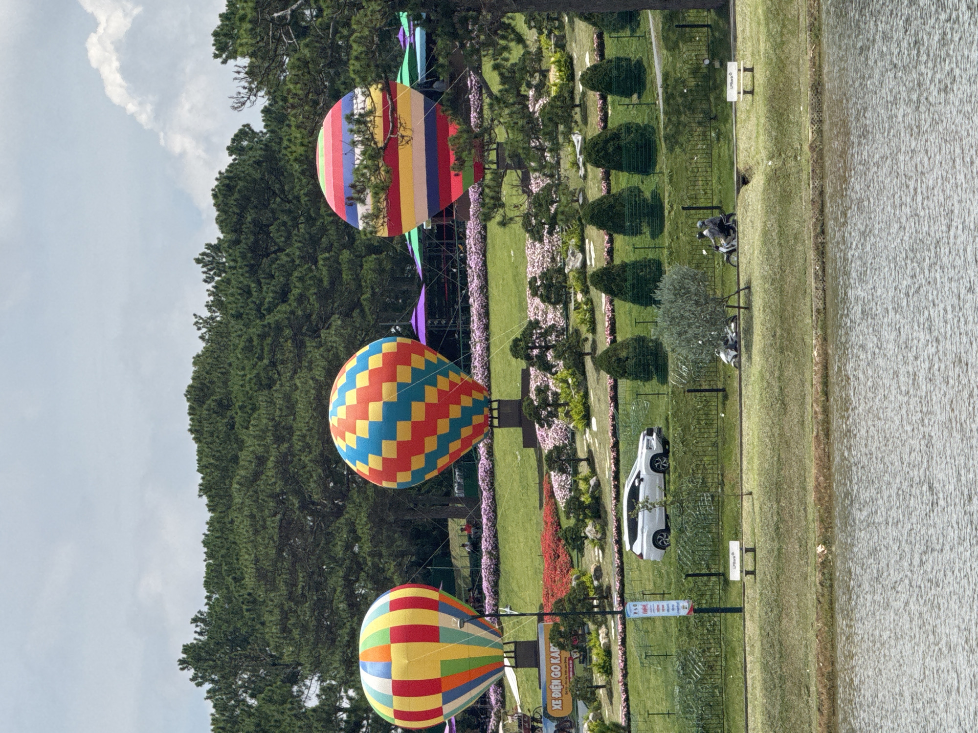 The image captures a scenic view of a park in Da Lat, Vietnam, featuring three large, vibrantly colored hot air balloons as the primary focal points. The balloons are patterned with geometric shapes and stripes in various hues of red, yellow, blue, and green. They are situated on raised platforms amidst a well-maintained garden with manicured greenery, flower beds, and small trees. In the mid-ground, a white SUV is parked on a grassy slope. Further back, several people are visible, some seated, and two individuals are riding motorcycles along a paved path. A body of water, likely a lake or pond, occupies the foreground of the image, reflecting the sky and surrounding environment. The background is dominated by lush green pine trees, characteristic of Da Lat's mountainous landscape, under a partly cloudy sky. The overall impression is a cheerful and picturesque outdoor recreational area.