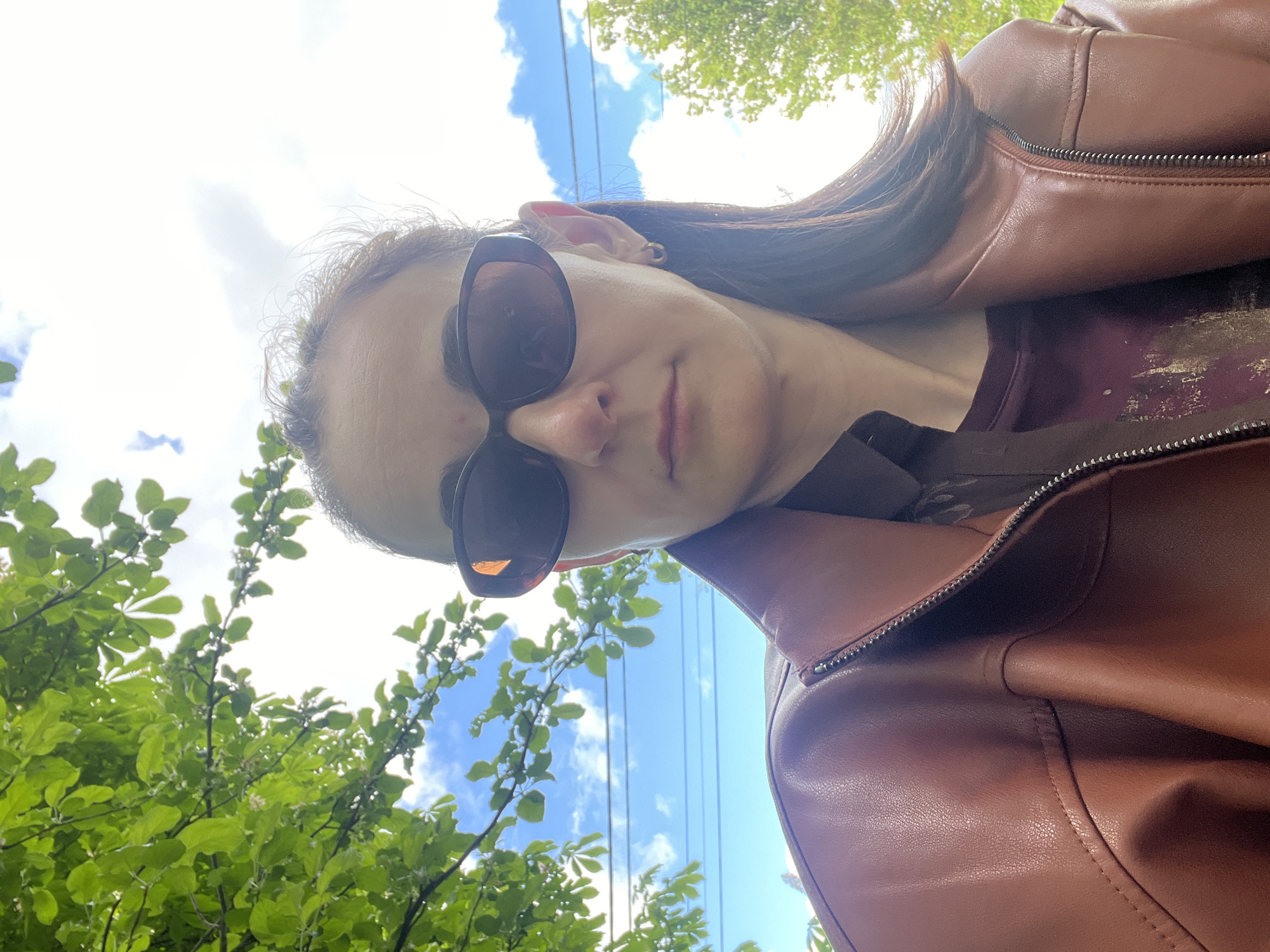 A selfie of a fair-skinned person wearing dark sunglasses and a brown leather jacket. The person is positioned at an angle, with the camera pointed upwards towards a blue sky with some white clouds. Green foliage, possibly from a tree, is visible in the lower left corner of the frame. The background location is the city of Unknown, Unknown.