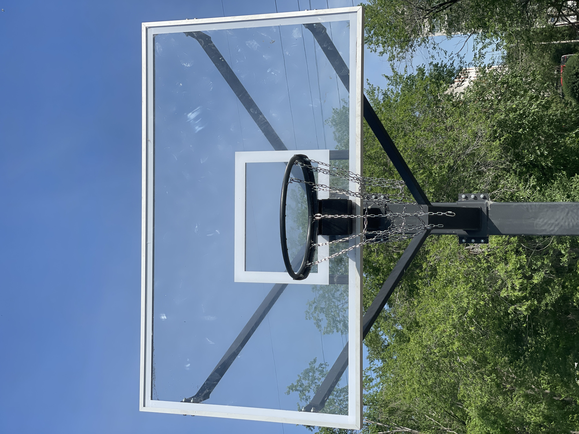 An outdoor basketball hoop with a clear backboard is shown against a bright blue sky. The backboard has a white rectangular outline around the hoop area. The hoop itself is black with a silver chain net. The structure supporting the backboard is black metal, and it is anchored to a gray pole. Lush green trees surround the base of the hoop, and some buildings can be seen in the background through the trees. The scene appears to be on a clear, sunny day.