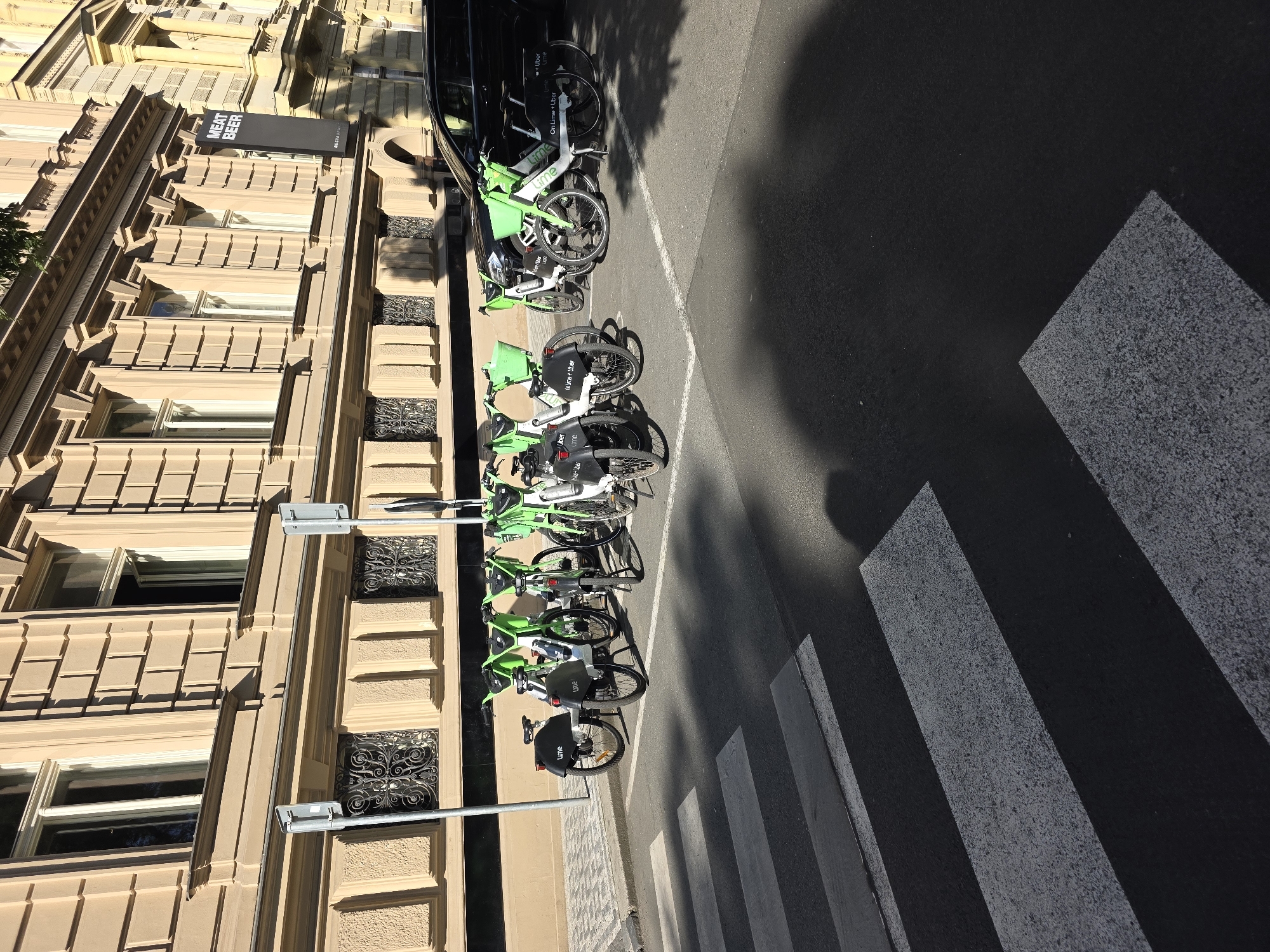 A sunny day scene captures a row of Lime rental bicycles parked on a cobblestone sidewalk next to a pedestrian crossing. The bicycles are predominantly green and white with black fenders and baskets. Behind the bicycles, a black SUV is parked in front of a building with ornate architectural details, including large windows and decorative stonework. A dark banner with white lettering that reads "MEAT BEER" is visible on the building. The ground features a pedestrian crosswalk with white stripes.