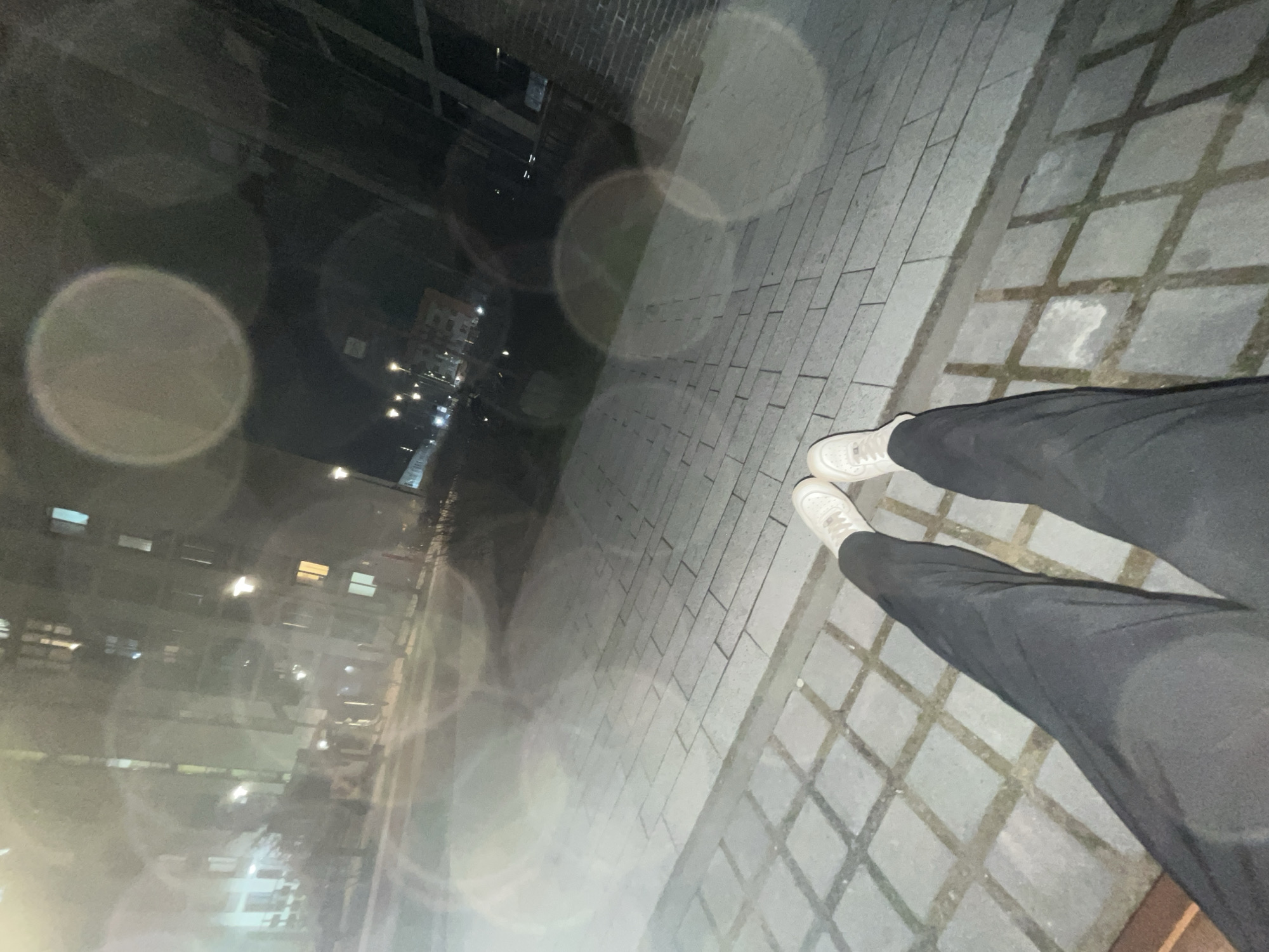 The image shows a person standing on a gray paved walkway in what appears to be an urban setting at night. The person is wearing dark pants and white sneakers. In the background, there are buildings, street lights, and possibly other structures. The scene is blurred with lens flare, and the angle is a low-angle point-of-view shot, possibly taken by the person standing there. The architecture is consistent with the city of Saint Petersburg, Russia.
