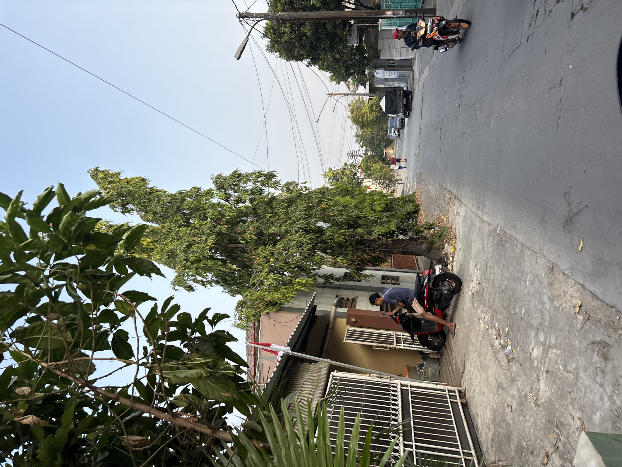 The image depicts a street scene in Bekasi, Indonesia. A young man wearing a blue t-shirt and light-colored shorts sits on a red and black scooter parked on the side of a paved road. To his left, a white metal gate leads to a building with a red and white Indonesian flag displayed on a flagpole. A tall, leafy tree stands behind the scooter and extends into the upper portion of the frame. The street is lined with houses, more trees, and utility poles with numerous wires. Several vehicles, including a black truck and two motorcycles, are visible on the road in the distance. The sky is clear and bright, suggesting daytime.