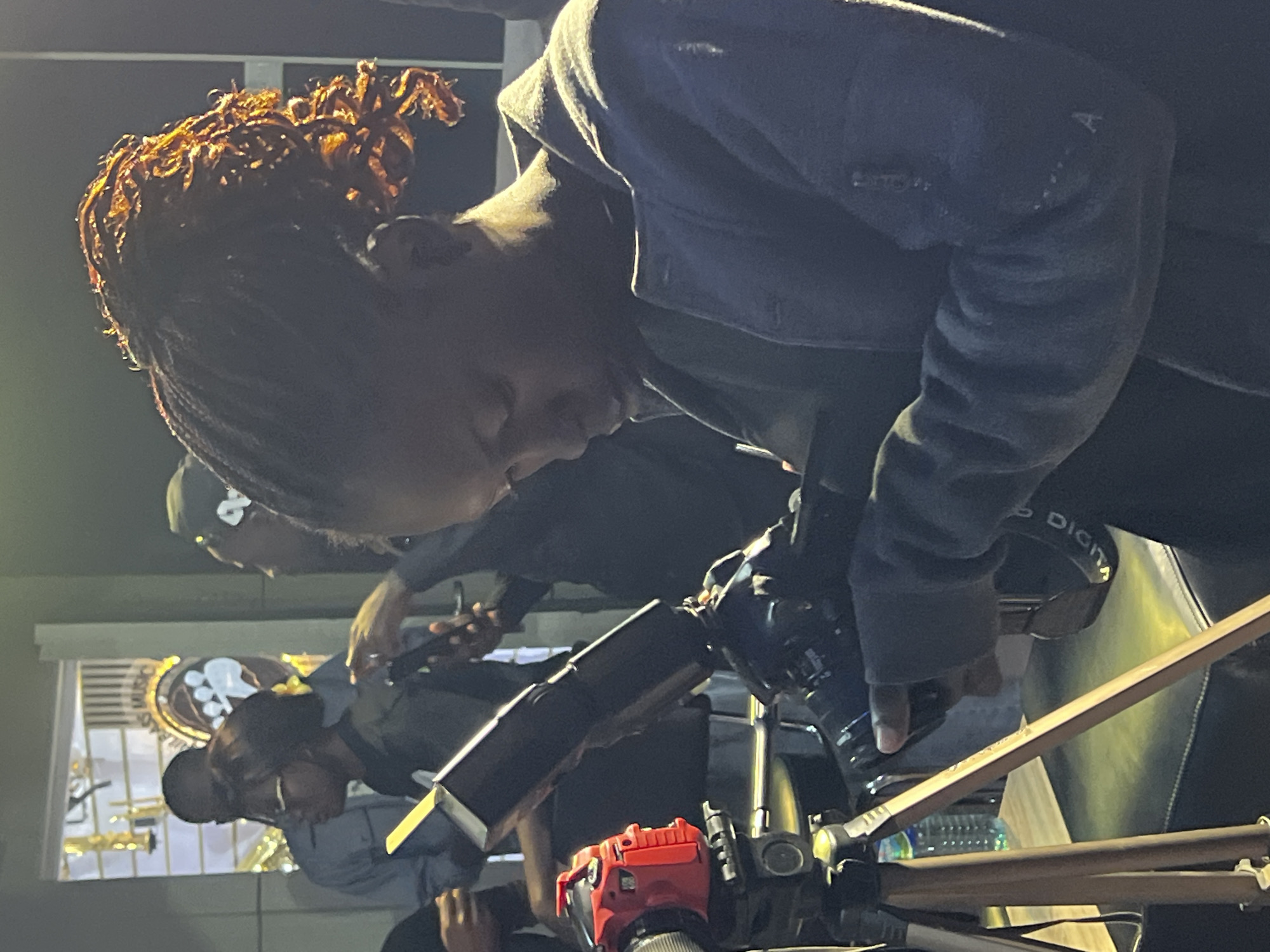 In what appears to be an indoor studio or recording space, likely in Jos, Nigeria, a young woman with braided hair styled in a high ponytail is captured in a close-up shot. She is intently focused on operating a camera, her hands manipulating the equipment. A red camera is visible on a tripod in the foreground to her left. Several other individuals are present in the background, some partially obscured, suggesting a collaborative or filming environment. Behind them, a display of musical instruments, including trumpets, can be seen through a doorway, hinting at a music-related setting. The lighting appears to be artificial, with a warm glow highlighting the woman's hair and shoulders. The overall scene conveys a sense of professional activity related to media production.