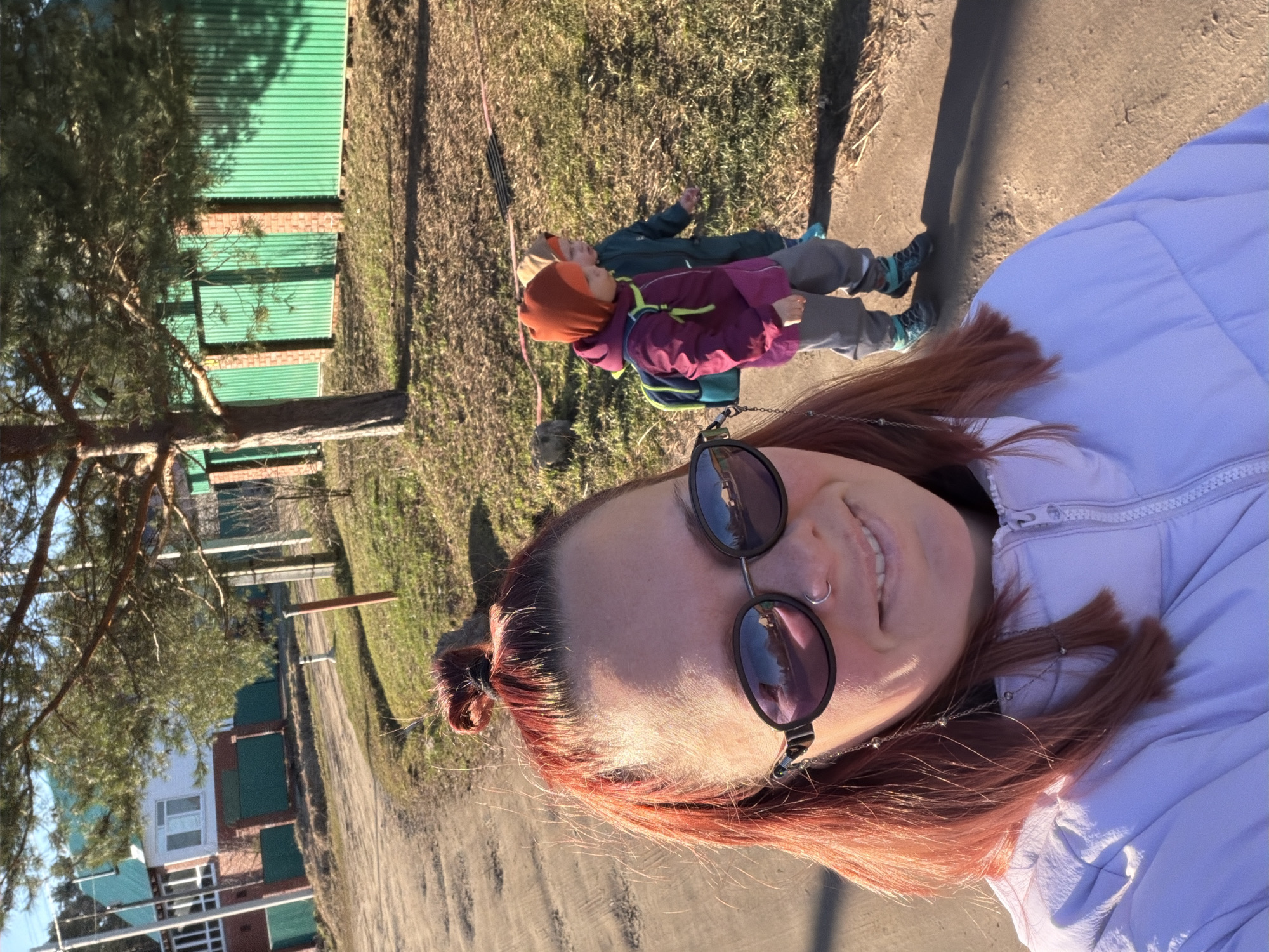The image is a selfie of a person with reddish-brown hair and sunglasses, wearing a light-colored jacket. In the background, two children are lying on a grassy area next to a concrete path. One child is wearing a red hat and a purple jacket with a green backpack, while the other is wearing a green jacket. Beyond the grassy area, there is a green metal fence and some trees. In the distance, a building can be seen. The location is likely Chaikovsky, Russia, based on the provided context.
