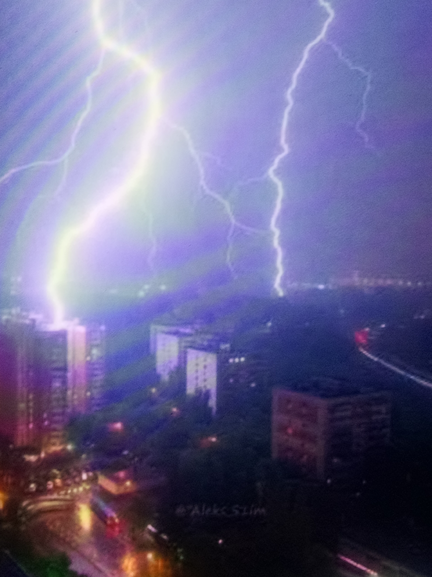 The image captures a dramatic night scene in an urban environment, likely Unknown, Unknown, during a thunderstorm. The sky is illuminated by multiple, powerful lightning strikes that cast an ethereal purple and green glow over the city. In the foreground, tall apartment buildings stand starkly against the stormy sky. Their windows show some signs of interior lighting. Below the buildings, a wet street is visible, reflecting the city lights and the lights of a passing bus or tram, suggesting recent rain or active precipitation. The overall atmosphere is one of intense natural power meeting the man-made landscape. The image appears to have a watermark on the lower right which reads "@Aleks_Slim".
