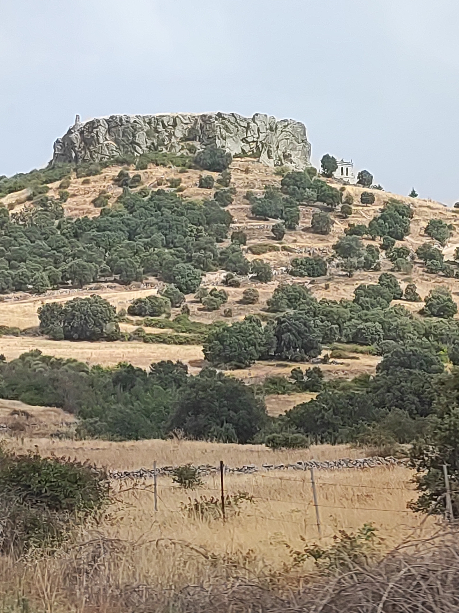The image captures a dry, hilly landscape dotted with trees, under a pale, overcast sky. Dominating the upper portion of the frame is a rocky summit, distinguished by its rugged, weathered appearance and a thin, upright structure atop its peak. To the right of this rocky outcrop, a small, white building with a dark roof and two prominent arched openings is situated on the hillside. The terrain below the summit is covered in dry, yellowish grass and scattered green trees. In the foreground, a rustic fence made of wire and wooden posts divides a field of dry, golden grass. The overall scene suggests a rural, possibly arid environment.