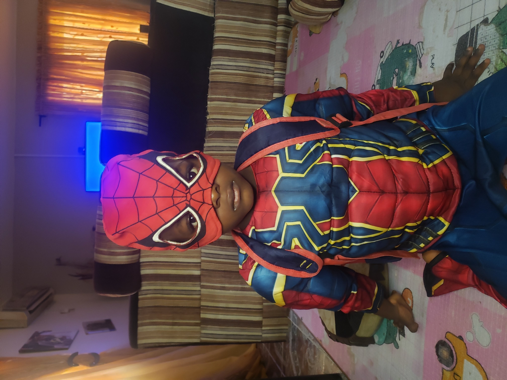 A young child wearing a full Spider-Man costume, including a mask that covers their head, is sitting on a pink and white mat with cartoon animal prints. The costume is red and blue with yellow accents. The child is looking directly at the camera, with their hands resting on the mat in front of them. The background shows a living room with striped cushions on a sofa, orange curtains, and framed pictures on the wall. An air conditioning unit is visible on the wall. The room appears to be in a residential setting in Narayi, Nigeria.