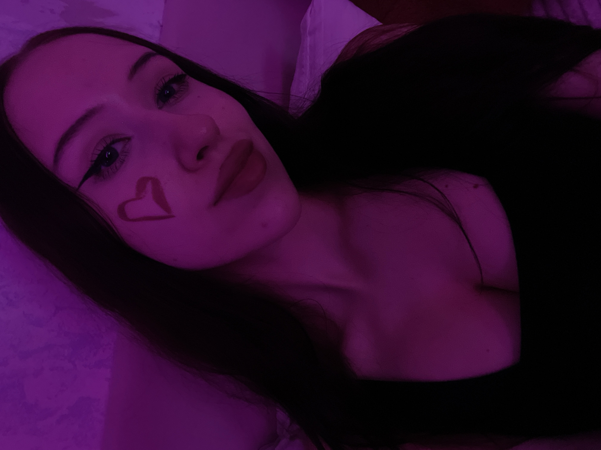 A young woman with long, dark hair is taking a selfie. She is wearing a black top. There is a heart shape drawn on her cheek. The lighting in the image is purple. The background appears to be a wall with a textured surface. There is a white pillow visible in the background. The image was taken indoors. There is no discernible location in Votkinsk, Russia, or any other specific city.