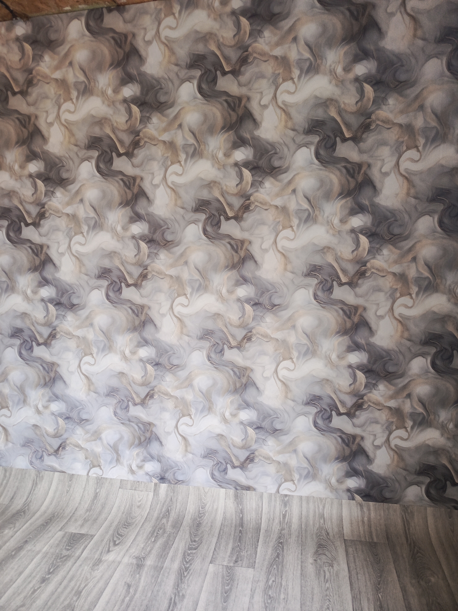 The image displays a corner of a room with a patterned wallpaper and a wooden floor. The wallpaper features a swirling, marbled design in shades of gray, white, and beige, creating an abstract, fluid appearance. The floor is made of light gray wooden planks with a visible wood grain pattern, laid at an angle. The lighting appears to be natural and diffused, suggesting an indoor setting during the day. There are no people or animals visible in the image. The scene is static and appears to be a still photograph of an unoccupied room. The location in the city of Meget, Russia, is not discernible from the visual elements present in the image itself.
